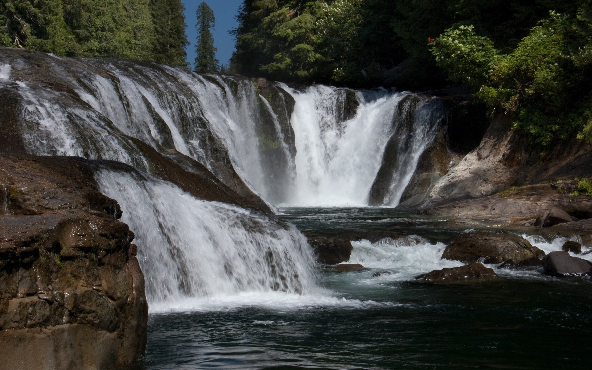 A wide waterfall cascades over rocks into a dark pool HD Wallpaper
