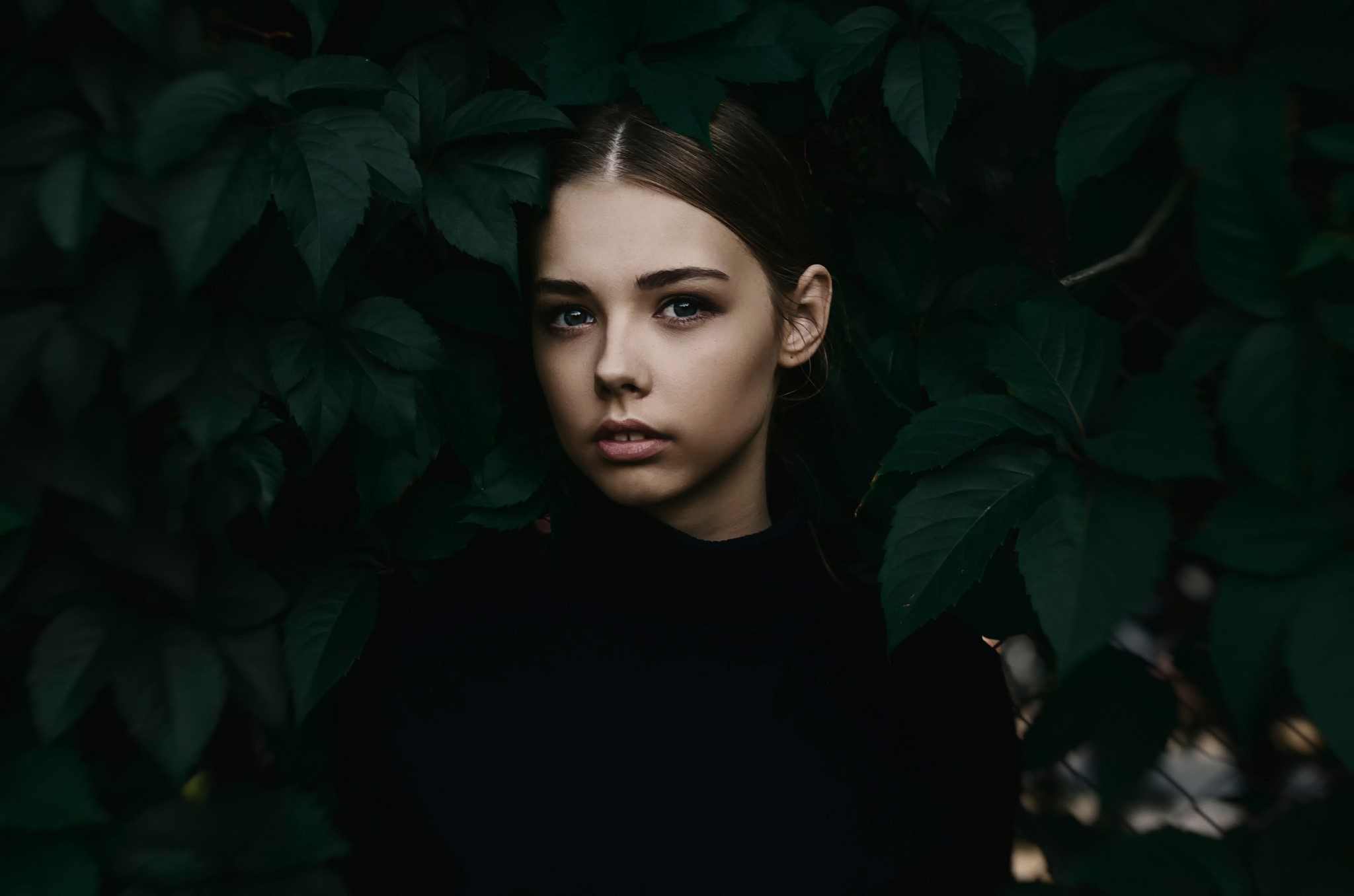 Young woman in black shirt surrounded by dark green foliage HD Wallpaper