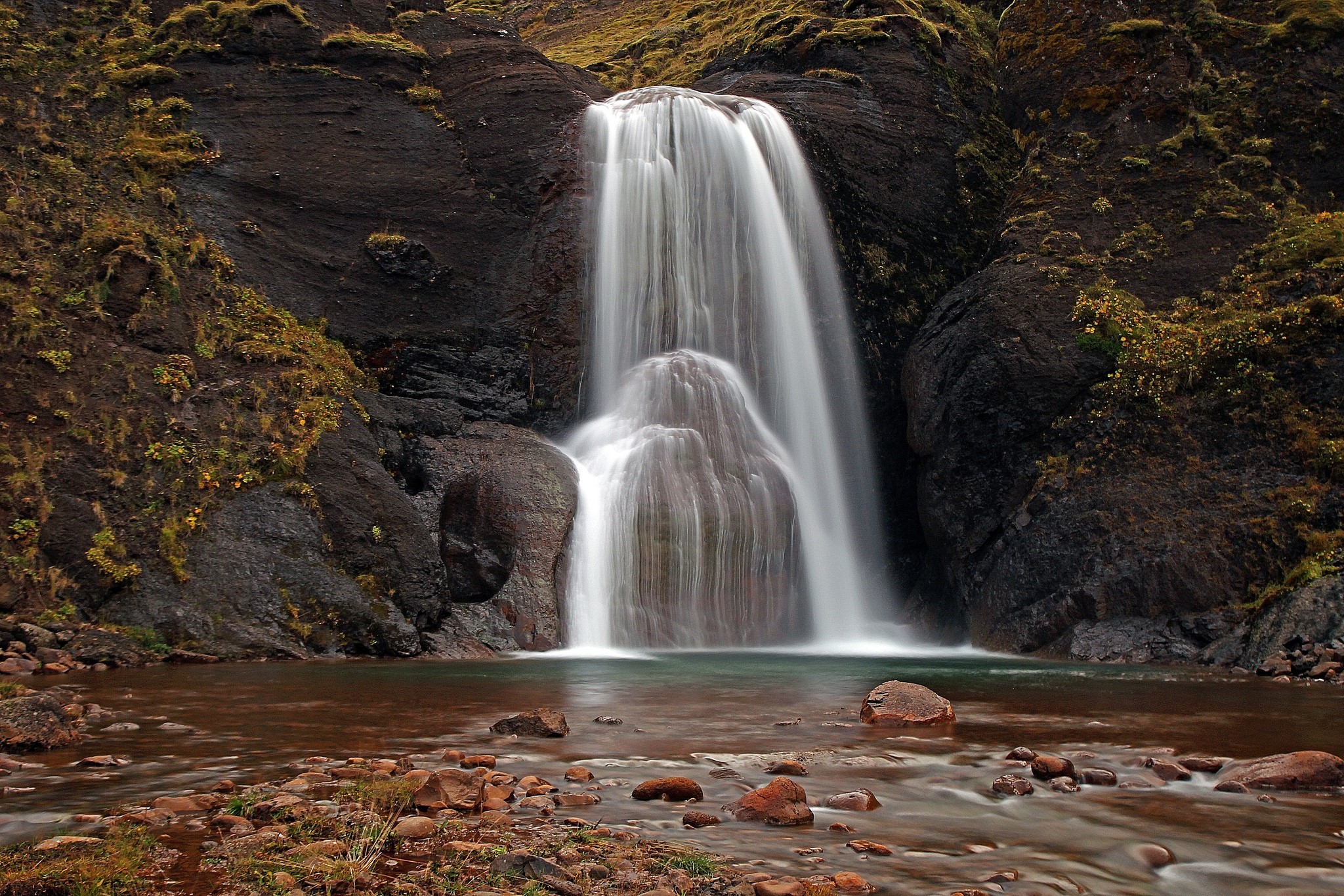 Waterfall cascading over dark rocks with stones in foreground HD Wallpaper