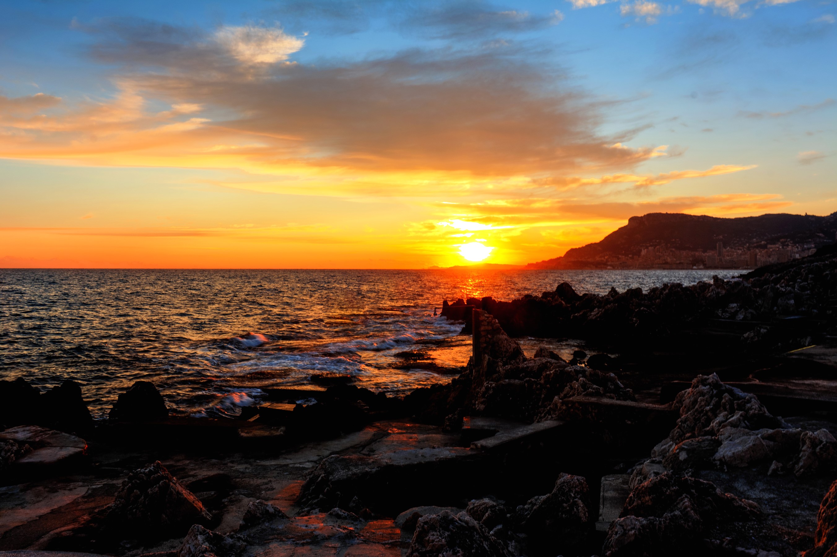 Sunset over the sea with rocky coast and distant town HD Wallpaper