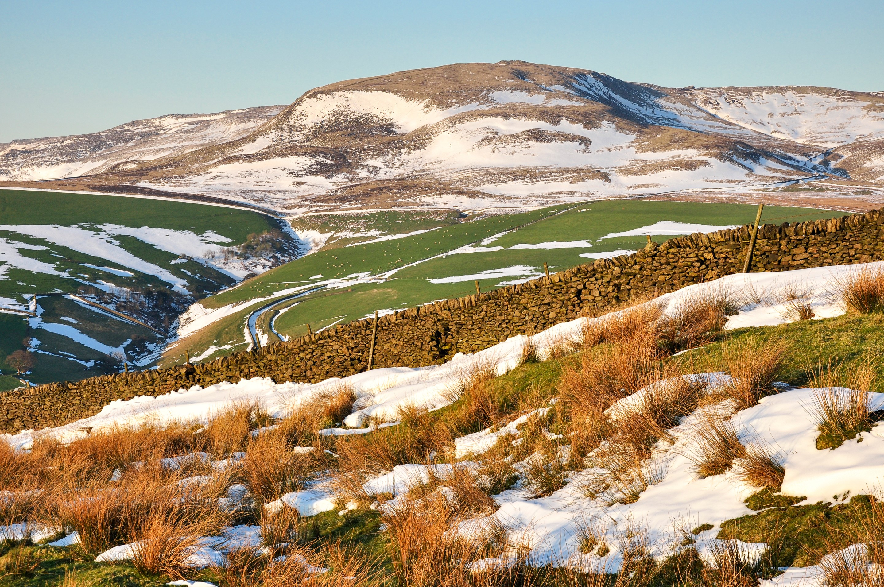 Snow-dusted mountains and green fields with stone wall HD Wallpaper