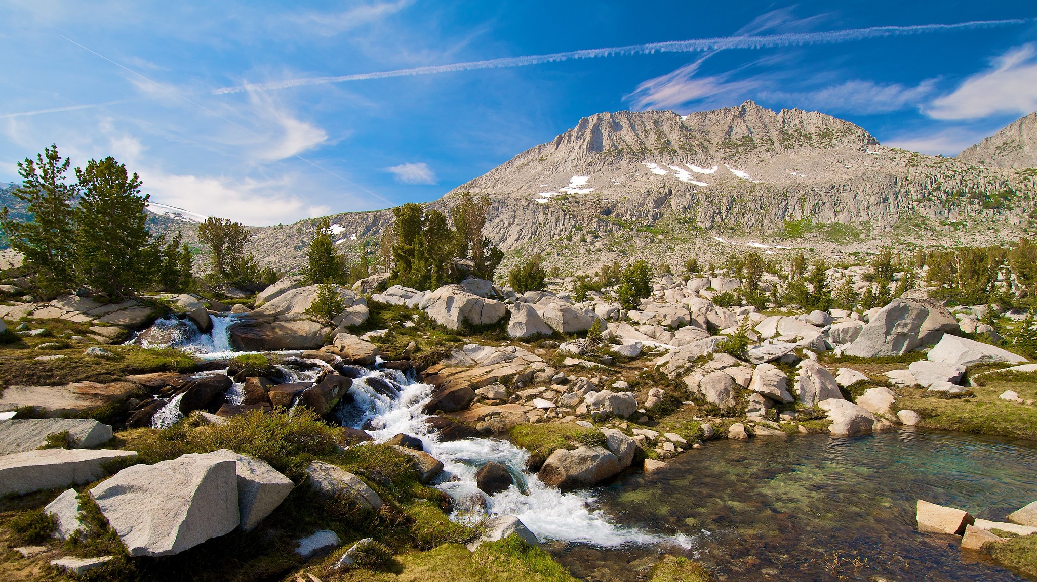 Mountain stream flowing through rocky terrain with trees and snow-capped peaks HD Wallpaper