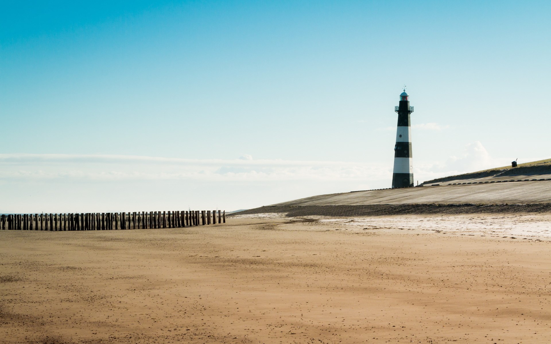 Lighthouse on a sandy beach with wooden posts HD Wallpaper