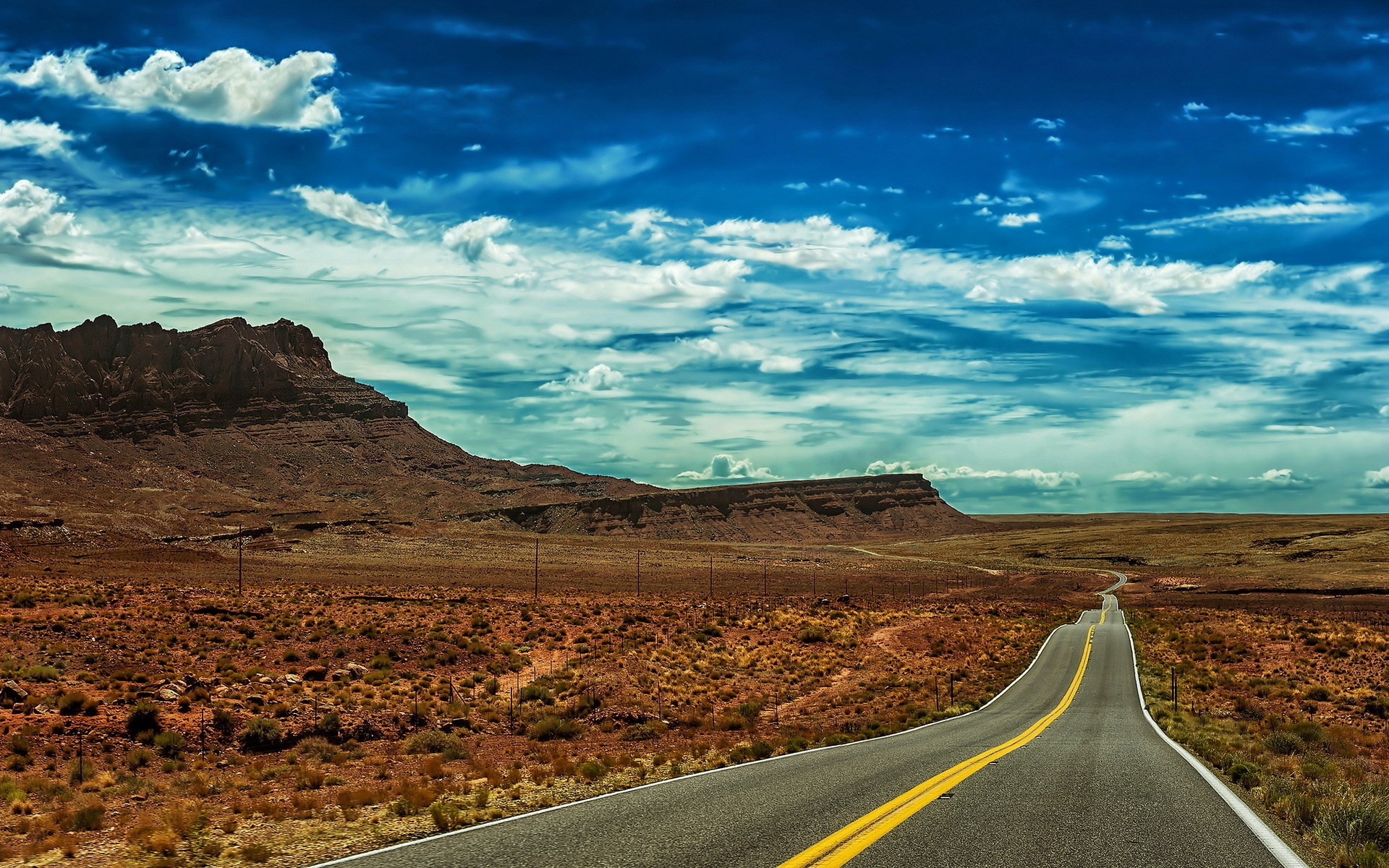 Desert road leading to mesas under a cloudy sky HD Wallpaper