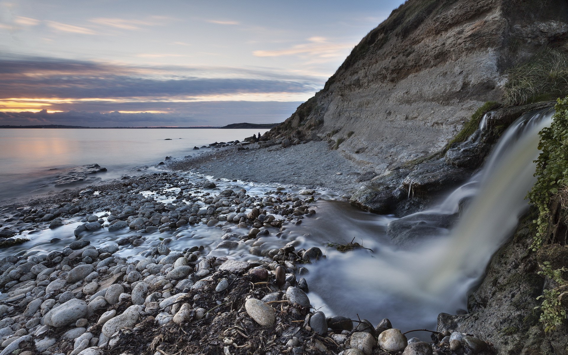Waterfall cascading onto a rocky shore at sunset HD Wallpaper