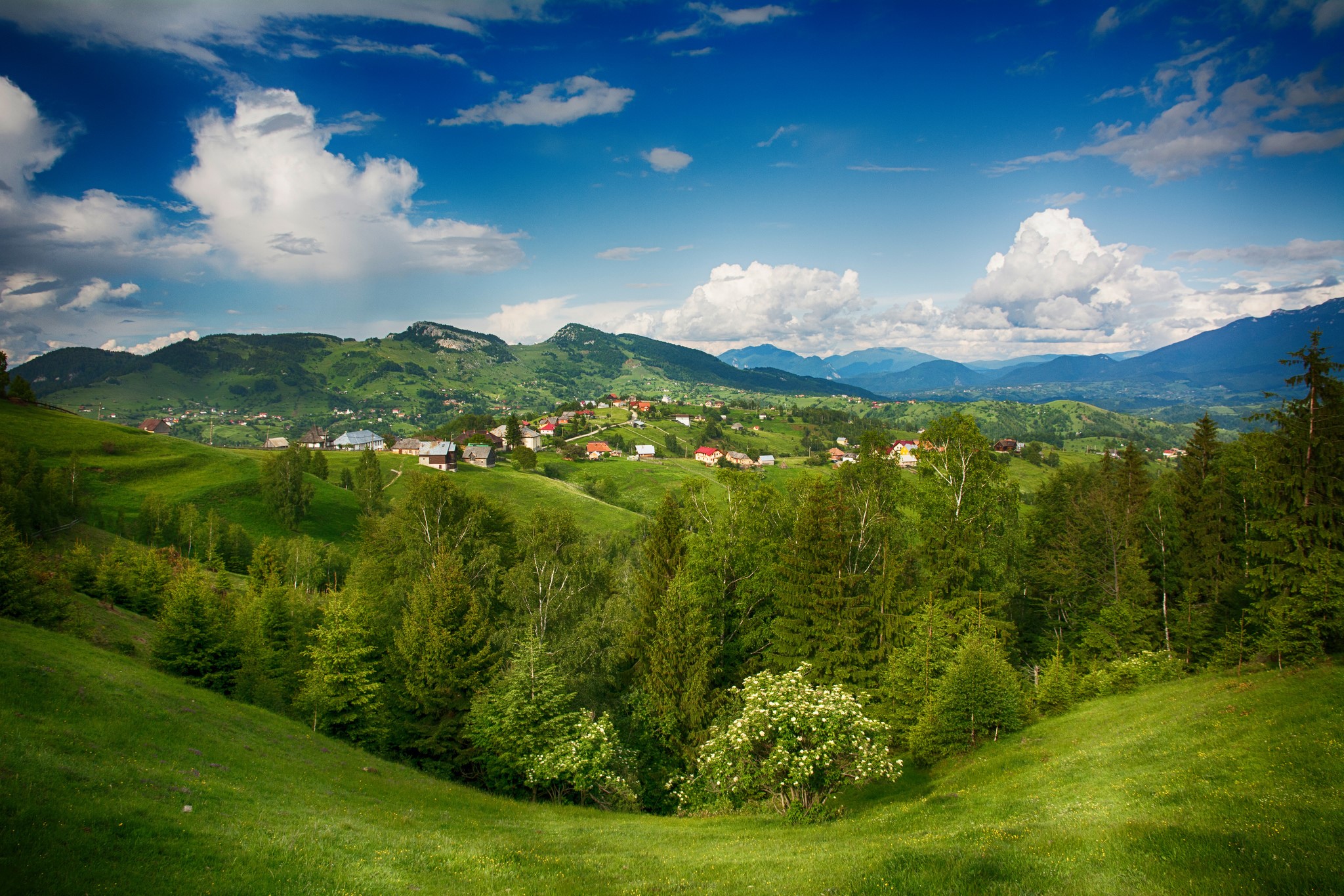 Green rolling hills with a village and mountains under a cloudy sky HD Wallpaper