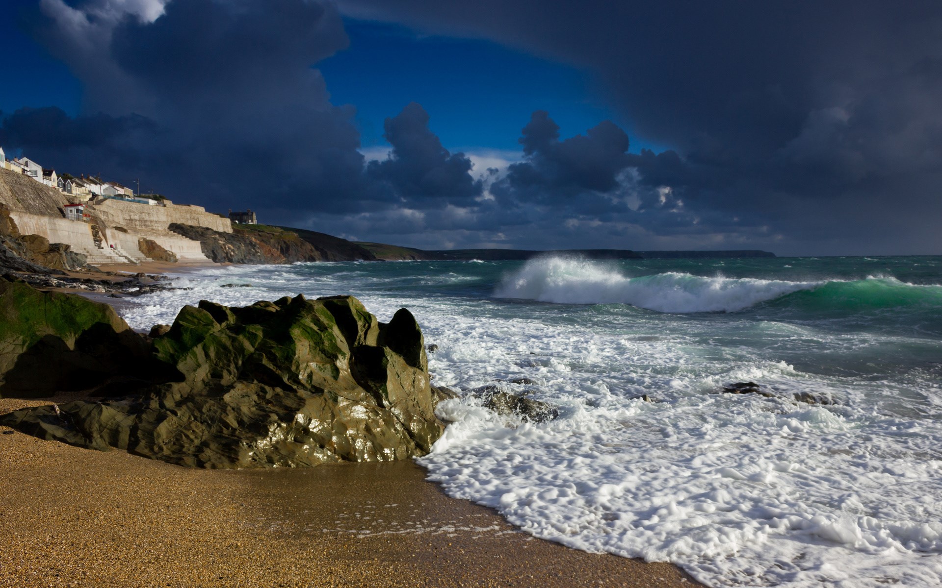 Stormy waves crash on a rocky beach with a village in the background. HD Wallpaper