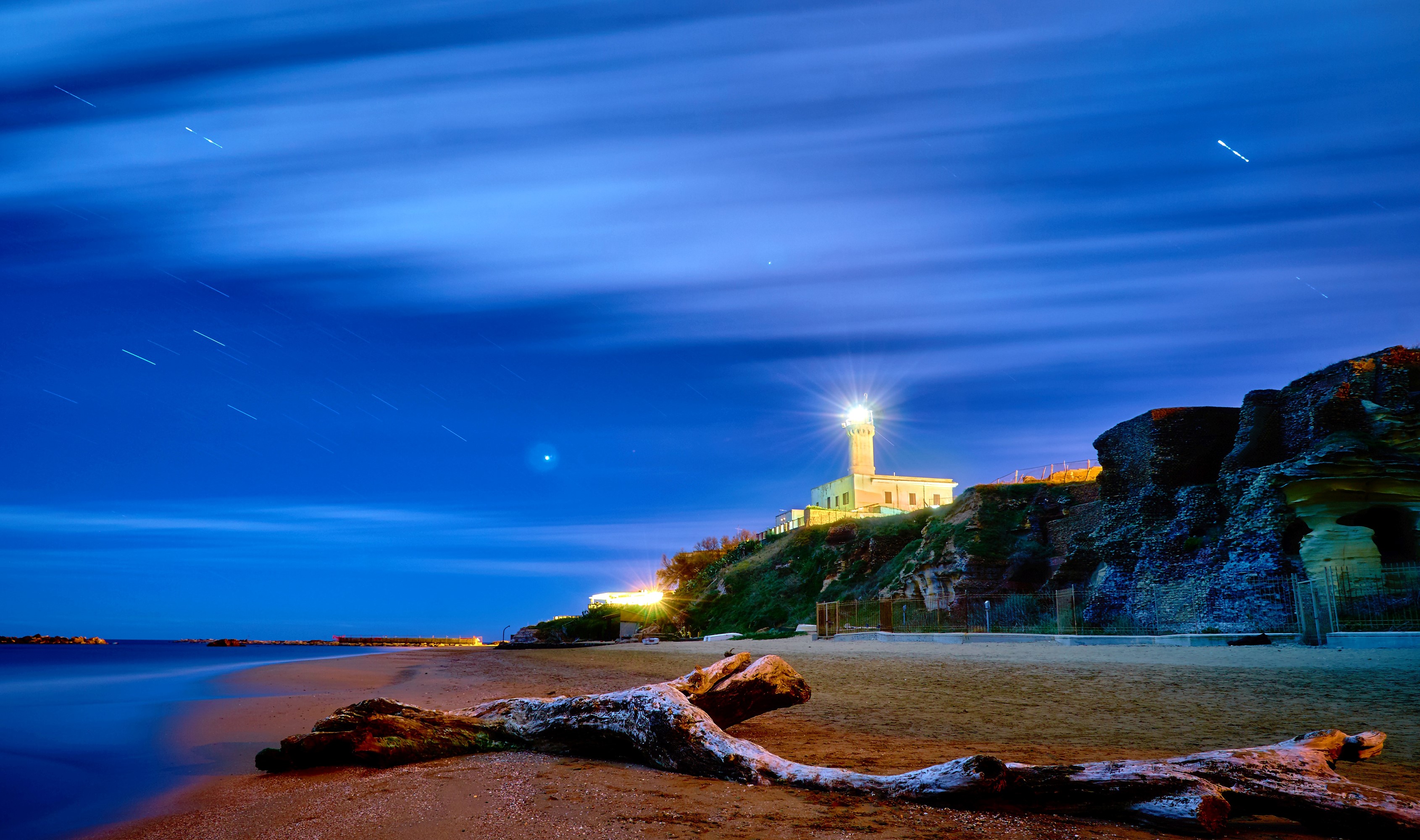 Lighthouse on a cliff at night with a driftwood log HD Wallpaper
