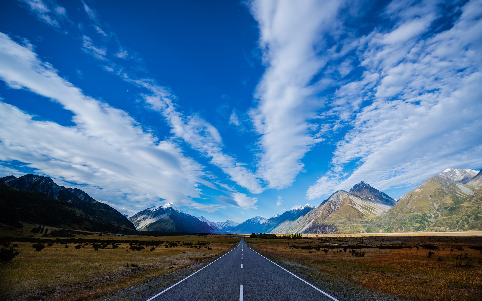 Empty road leading to snow-capped mountains under a cloudy sky HD Wallpaper