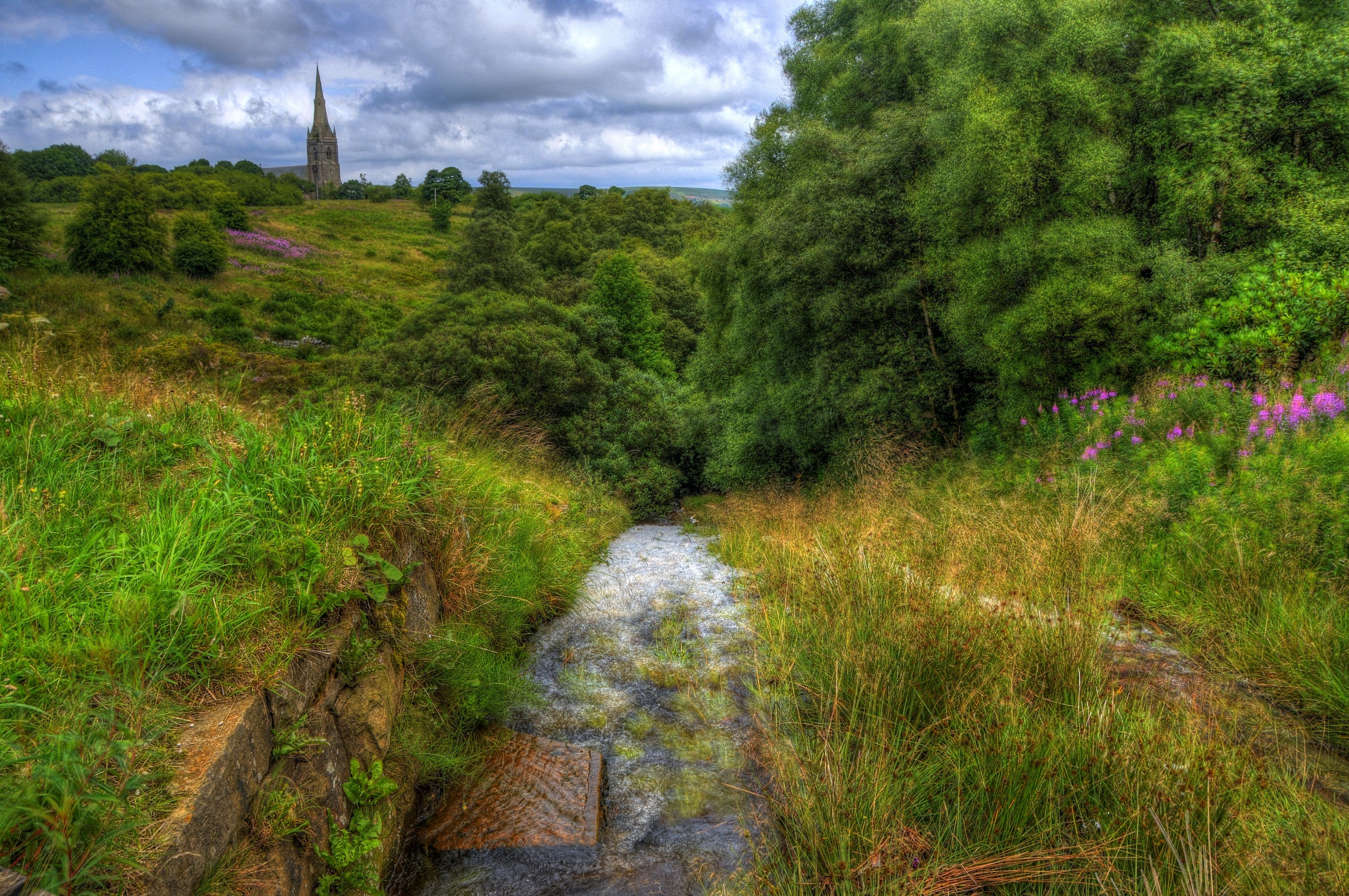 Stream flowing through a green landscape with a church spire in the distance HD Wallpaper