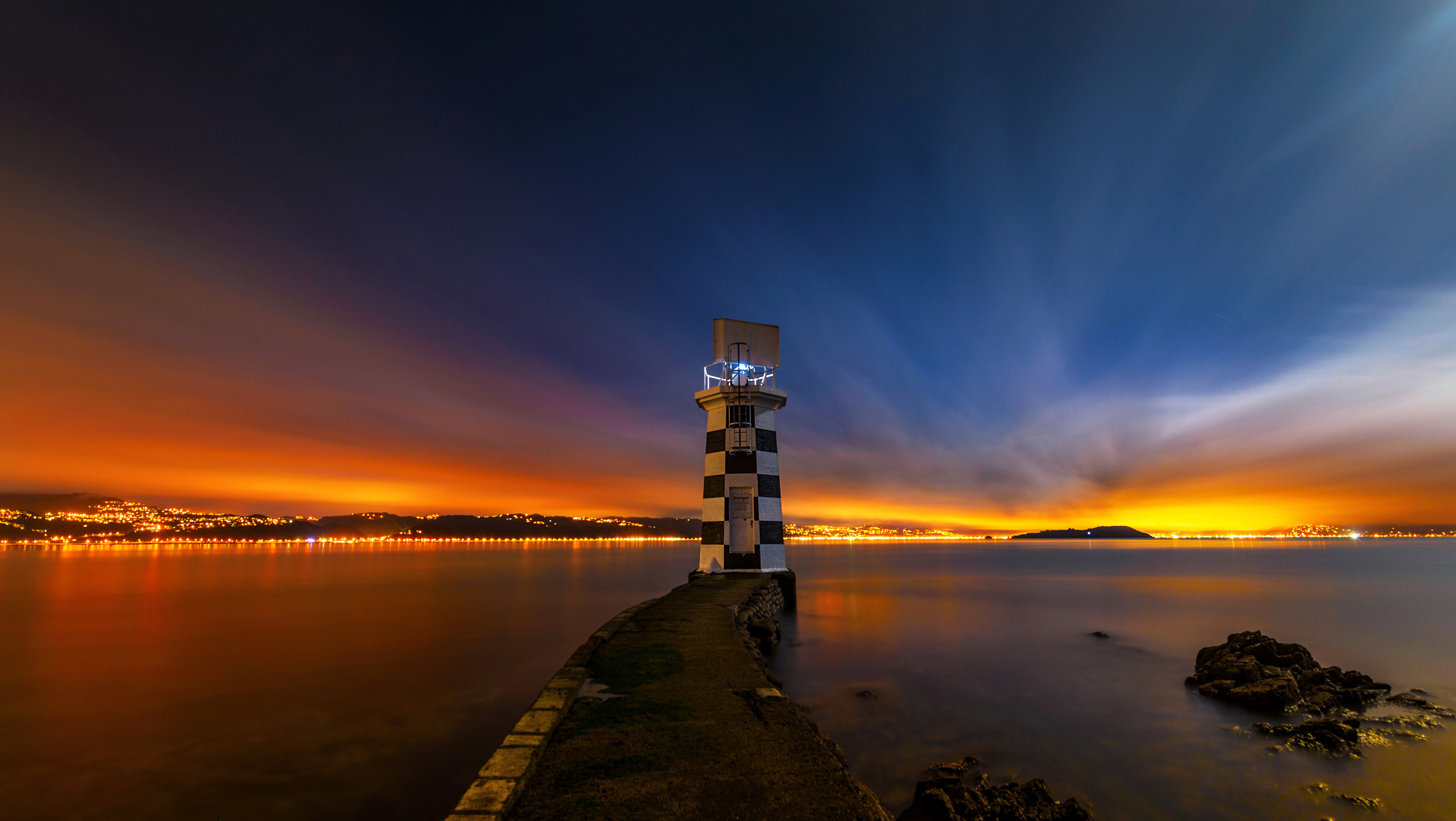 Lighthouse at night with city lights and sunset sky HD Wallpaper