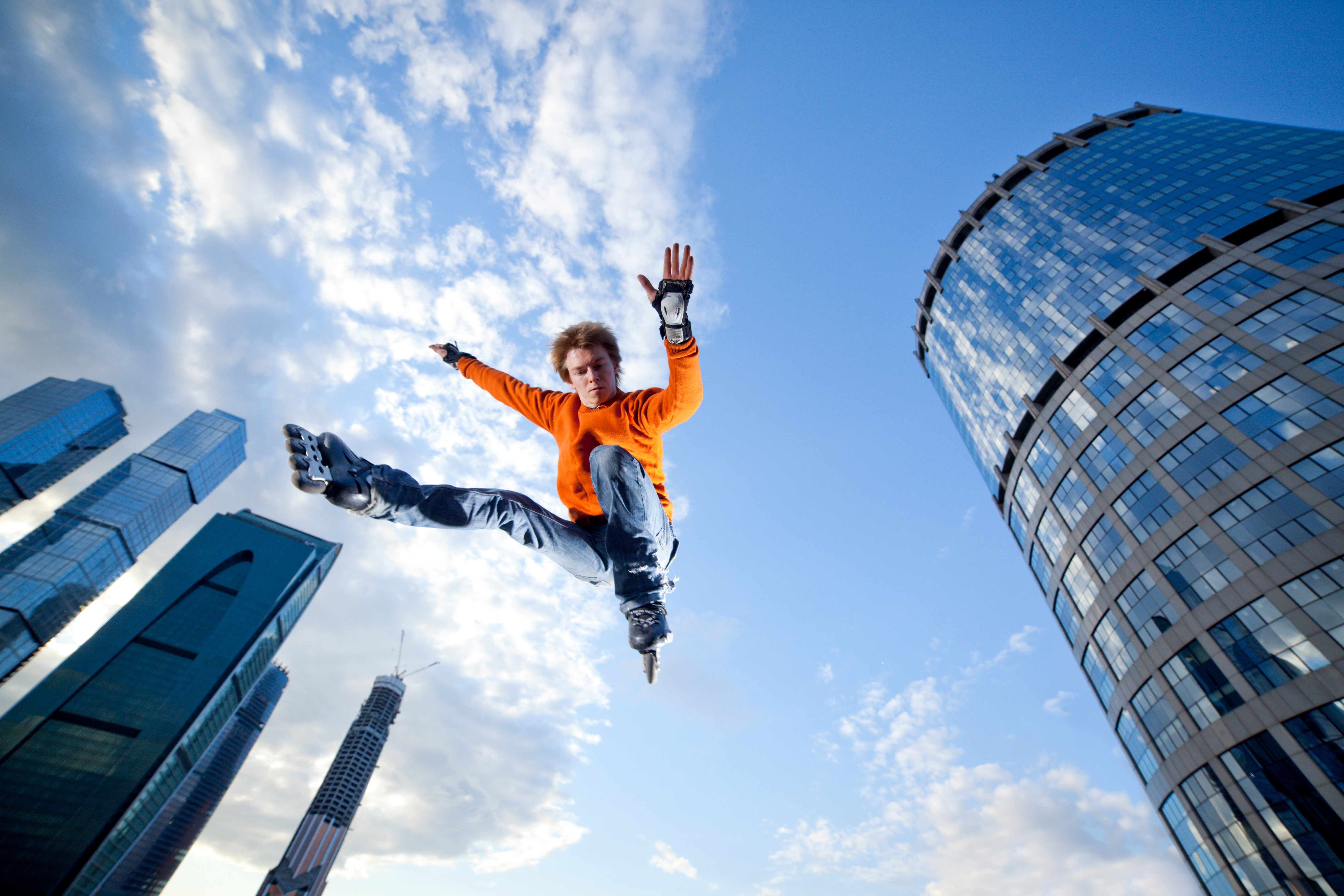 Man in orange sweater jumps on rollerblades with skyscrapers in background HD Wallpaper