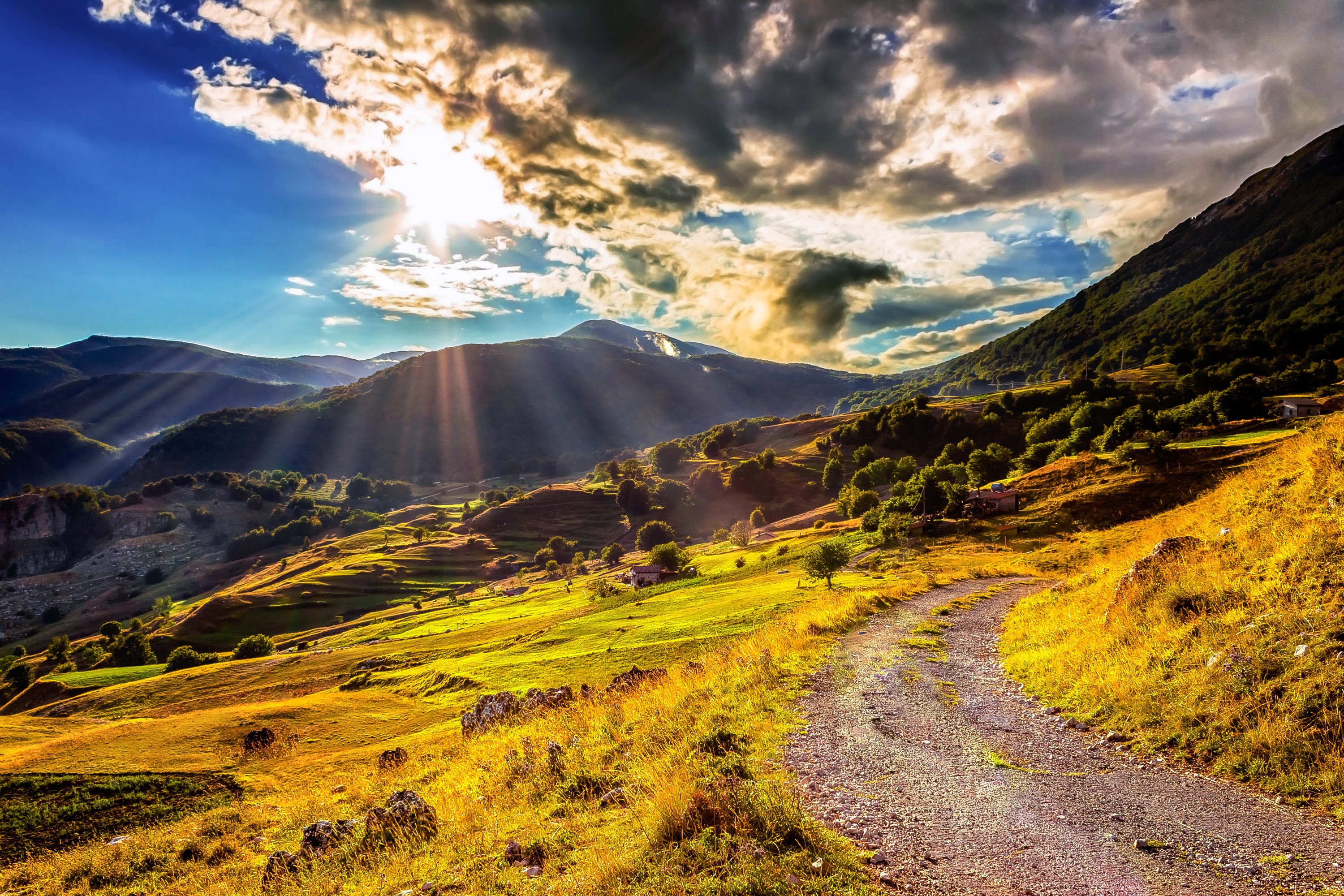 Sunlight beams over a mountain valley with a dirt road HD Wallpaper