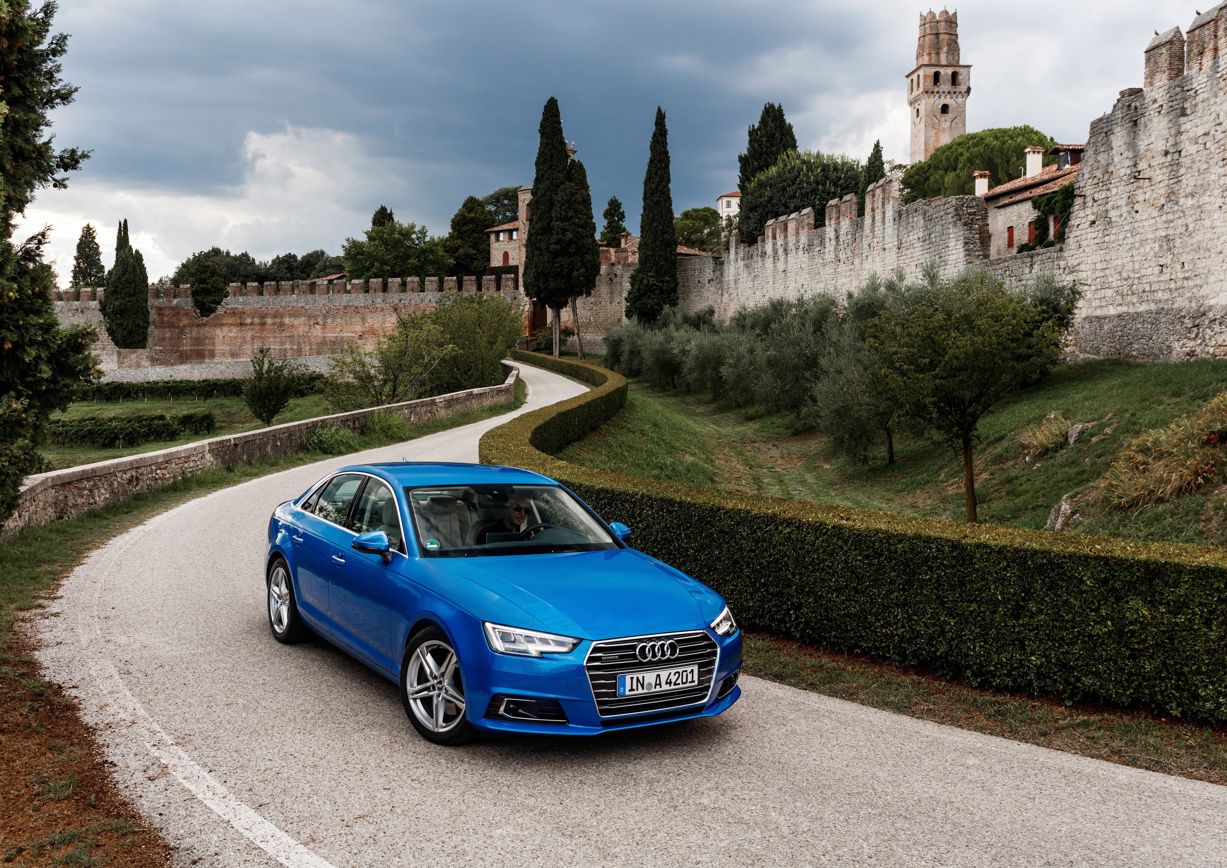 Blue Audi A4 driving on a road near a castle wall HD Wallpaper