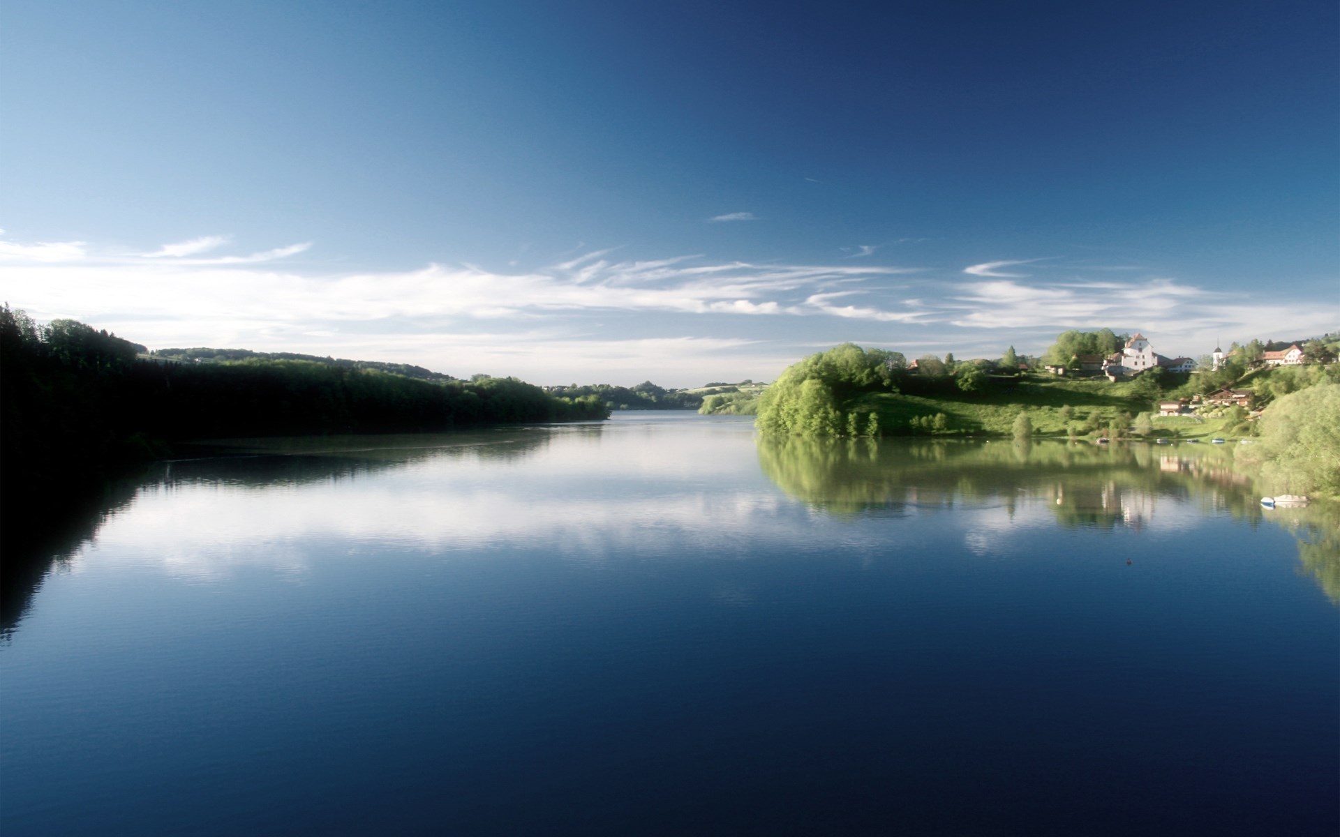 Calm lake reflecting trees and houses under a blue sky HD Wallpaper