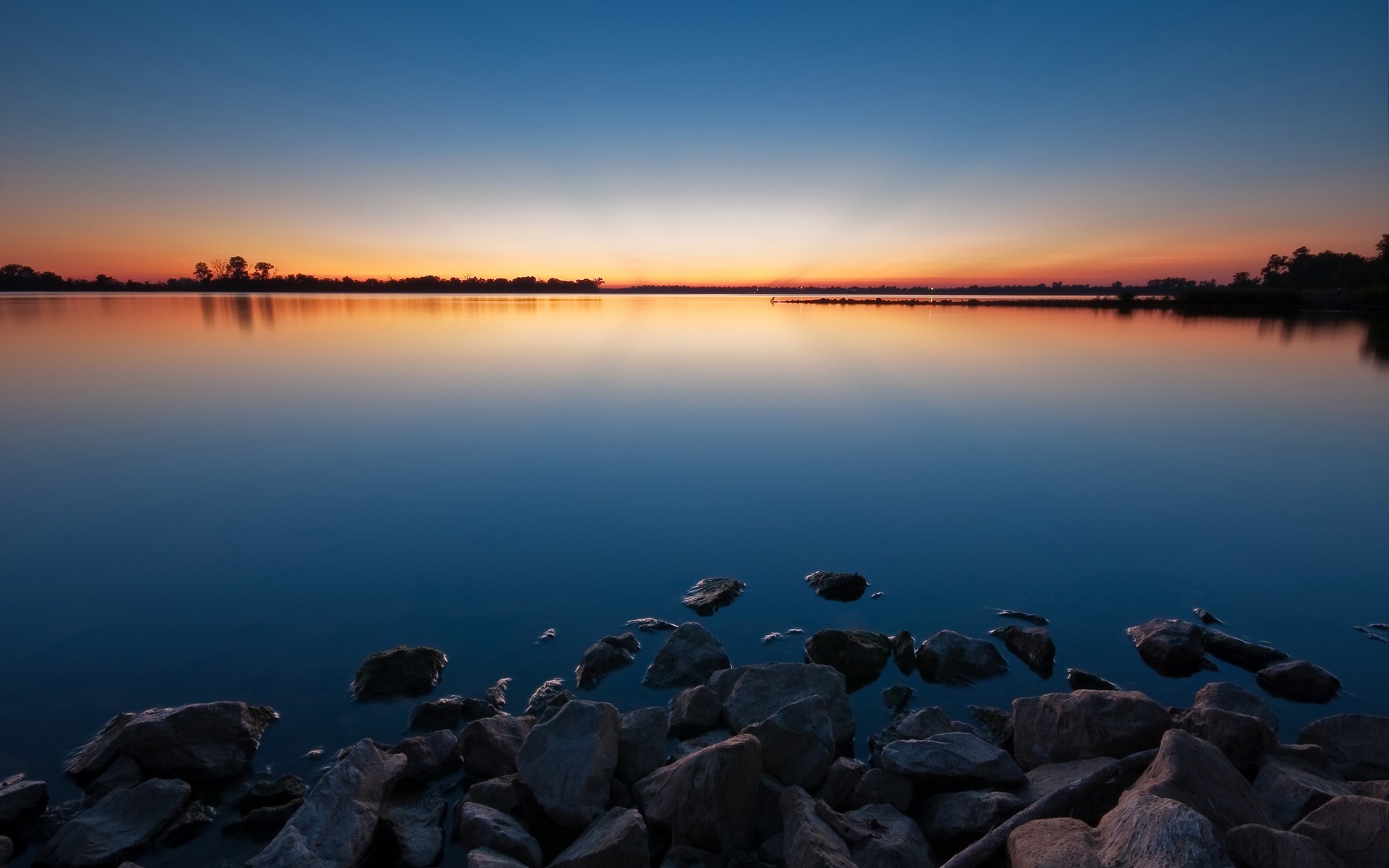 Calm lake at sunset with rocks in foreground HD Wallpaper