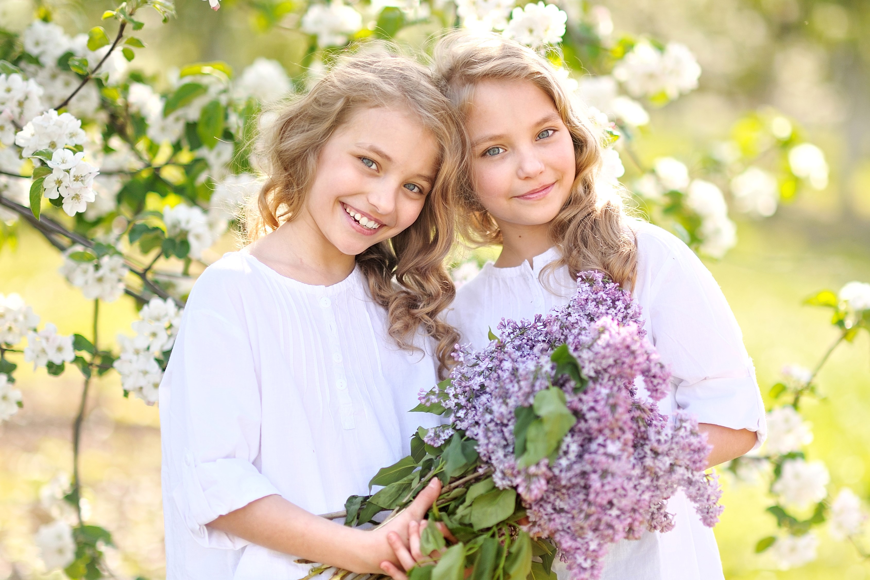 Two blonde girls holding lilacs in front of blooming tree HD Wallpaper