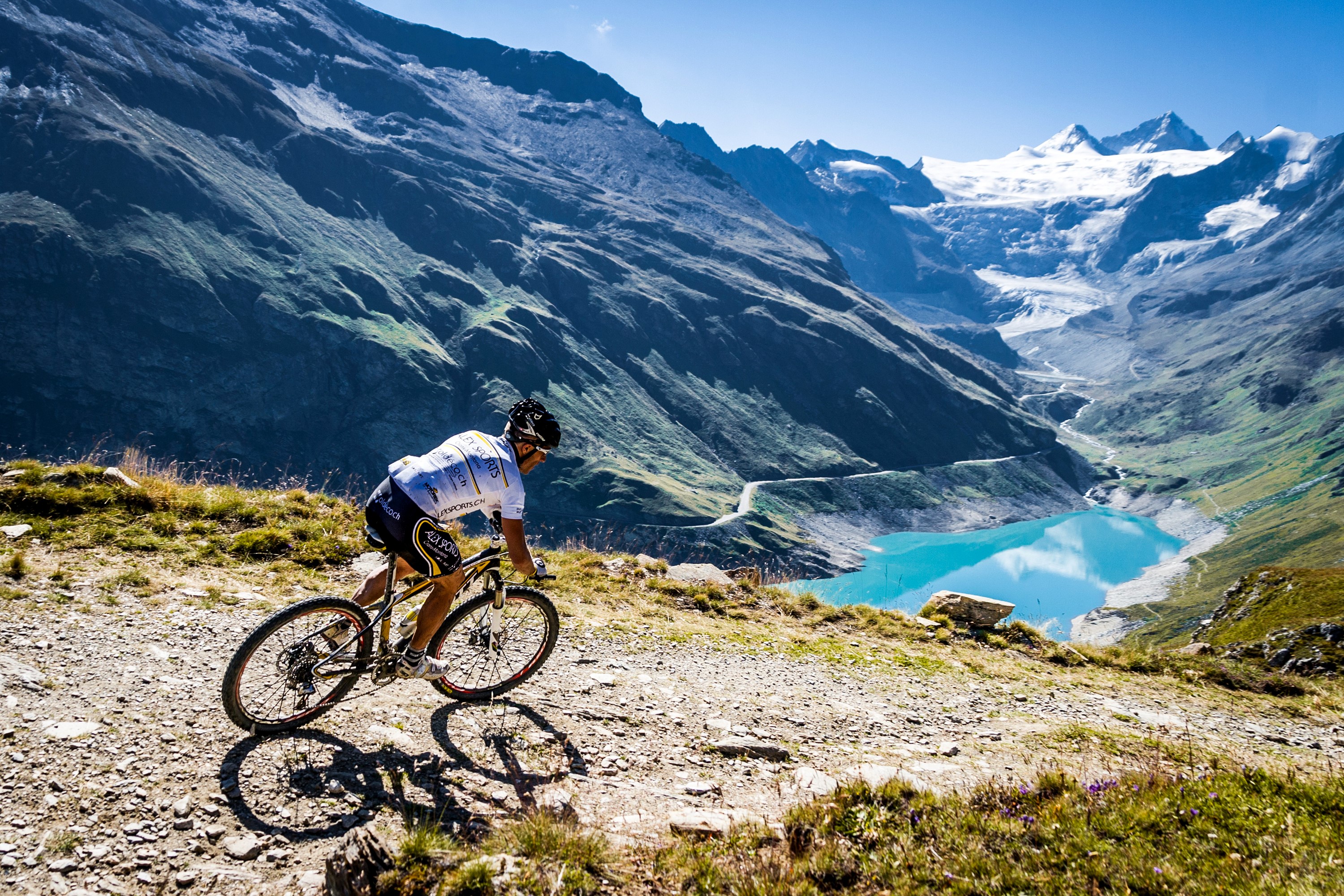 Man mountain biking near a turquoise lake with glaciers HD Wallpaper