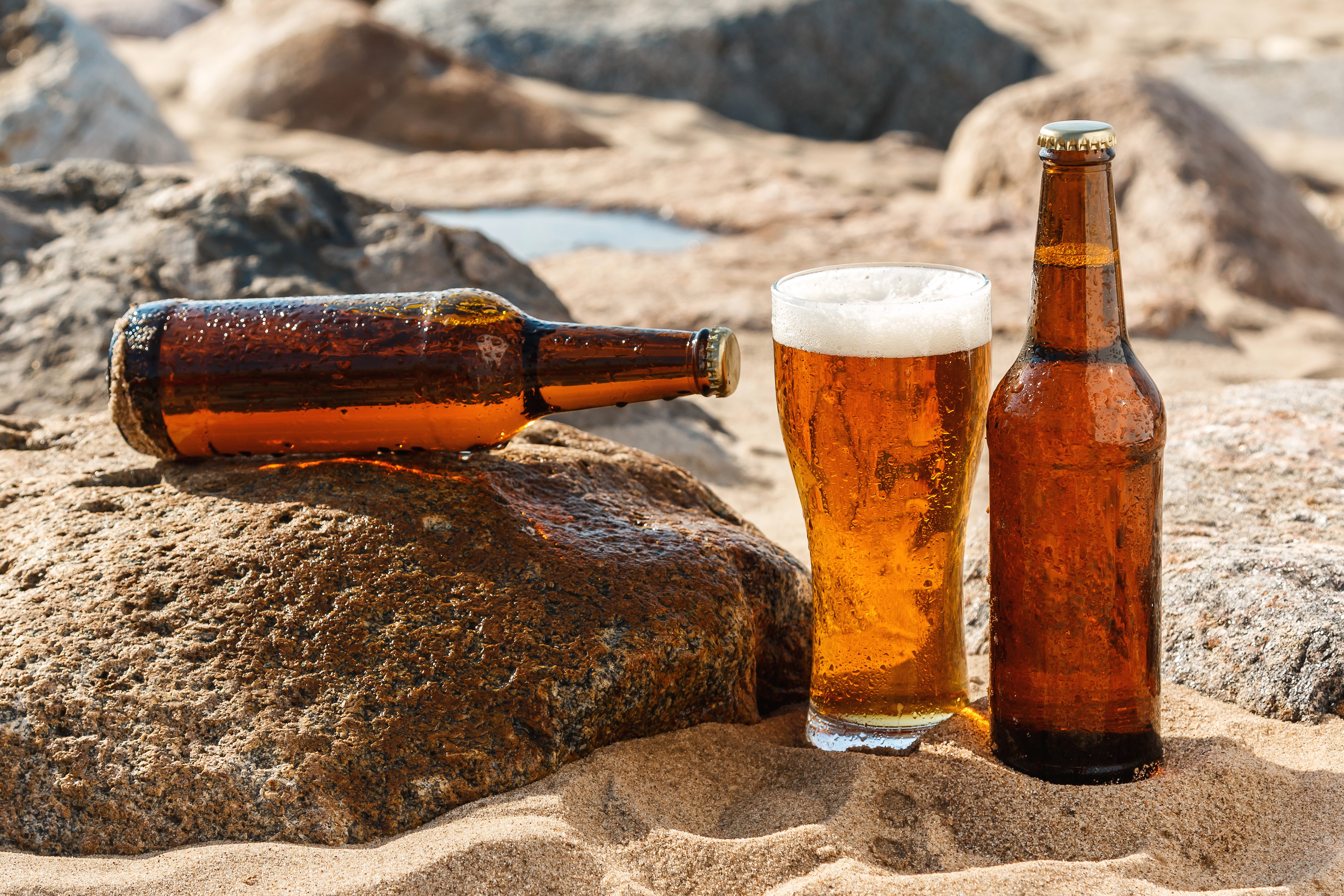 Beer bottles and glass on sand with rocks HD Wallpaper