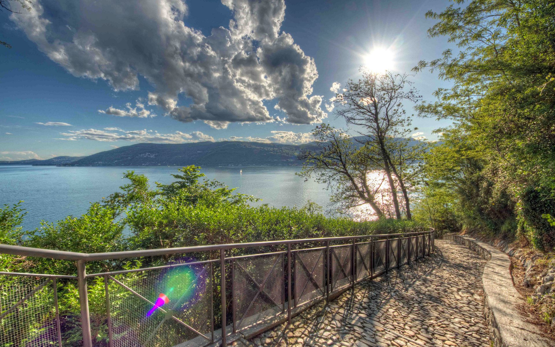 Cobblestone path along a lake with mountains and sun HD Wallpaper