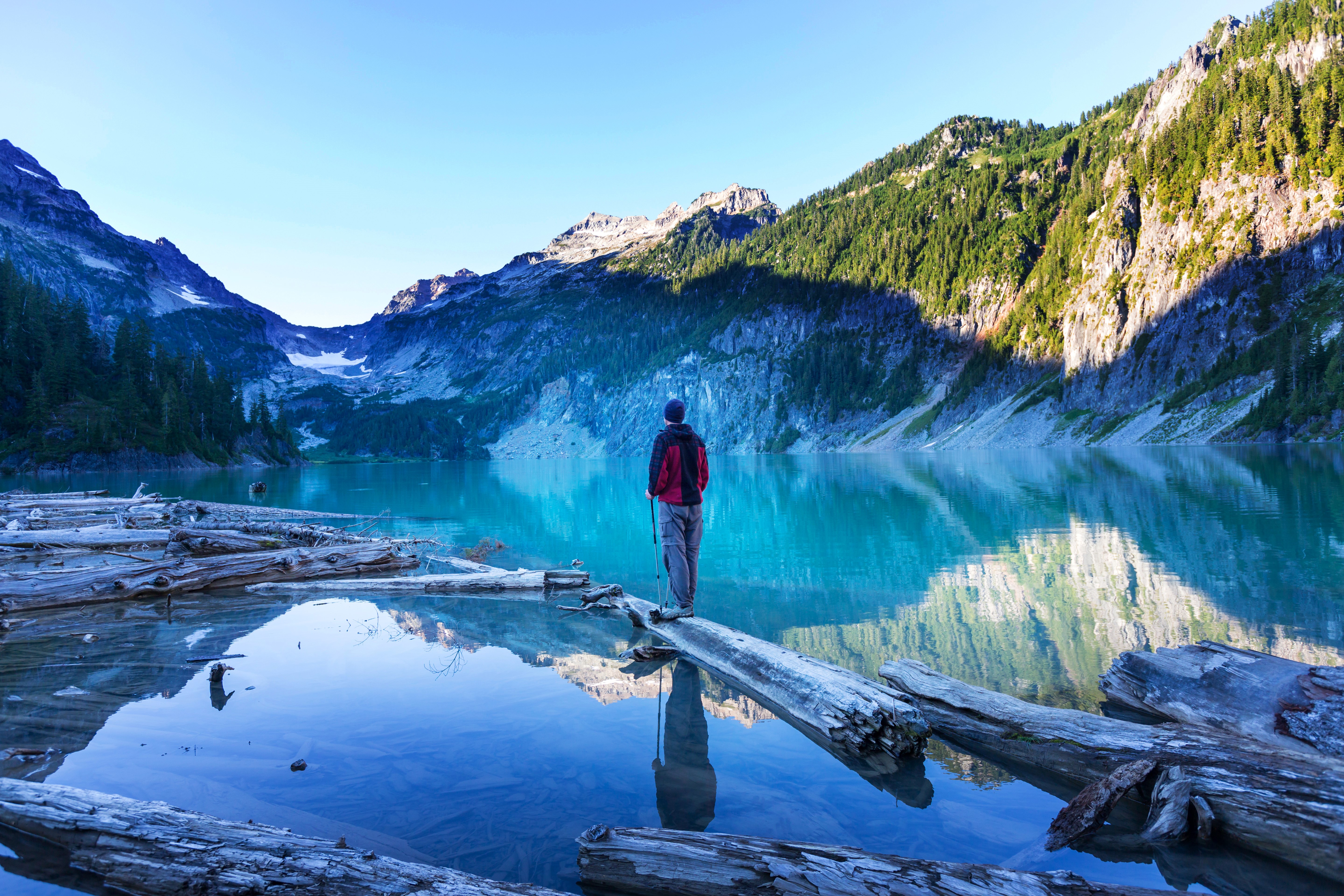 Man standing on logs in a lake with mountains HD Wallpaper