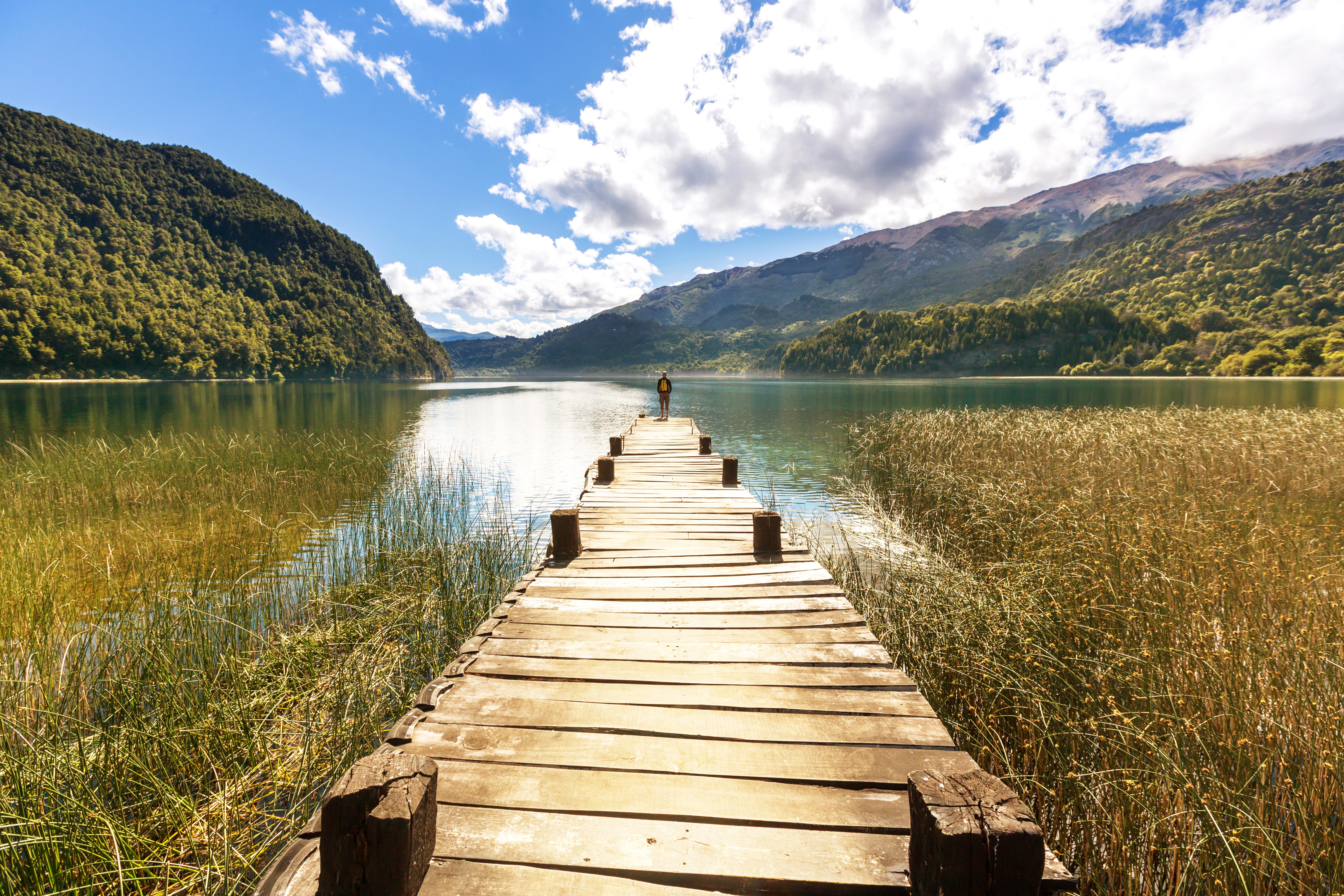Person on a wooden pier at a lake with mountains HD Wallpaper