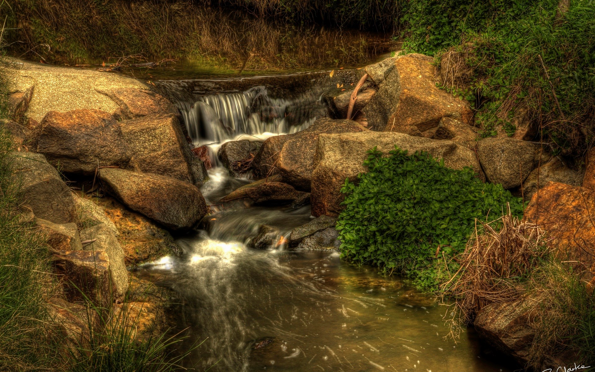 Small waterfall flowing over rocks with surrounding greenery HD Wallpaper