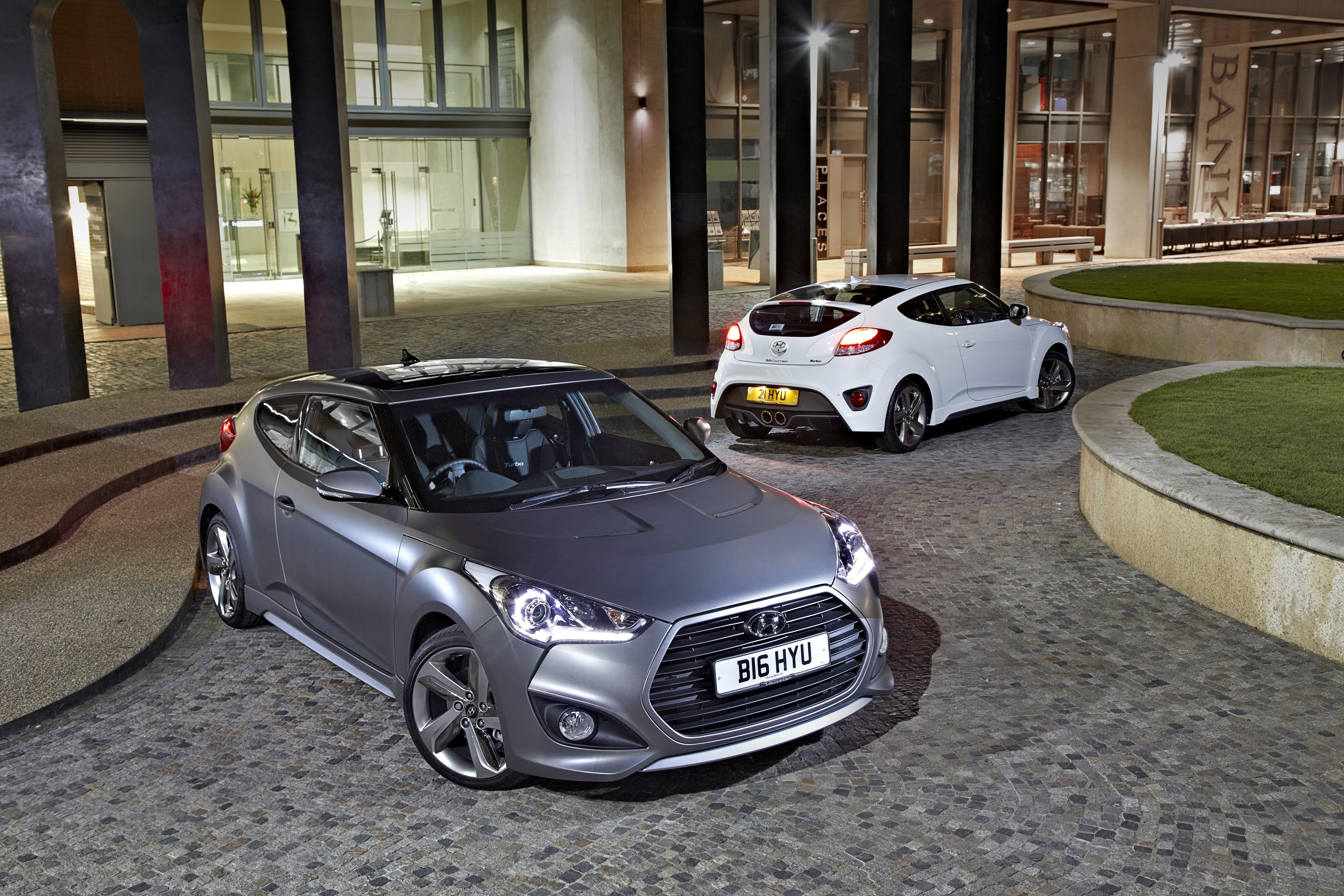Two Hyundai Veloster Turbo cars parked outside a modern building HD Wallpaper
