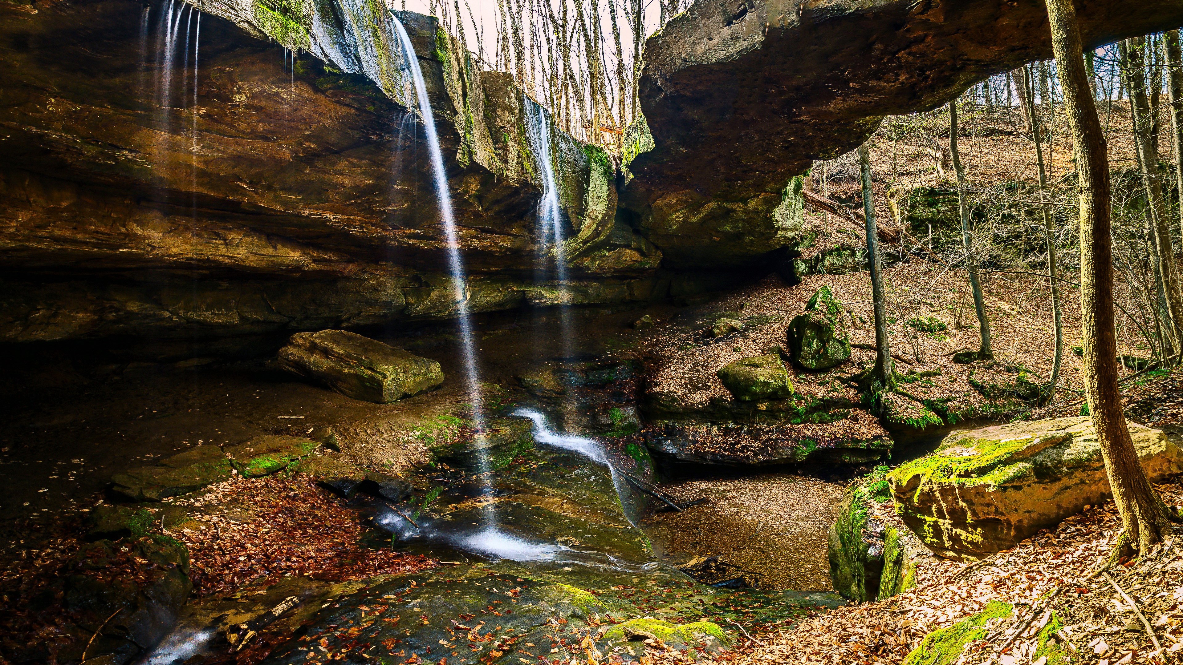 Waterfall flowing under a rock arch in a forest HD Wallpaper