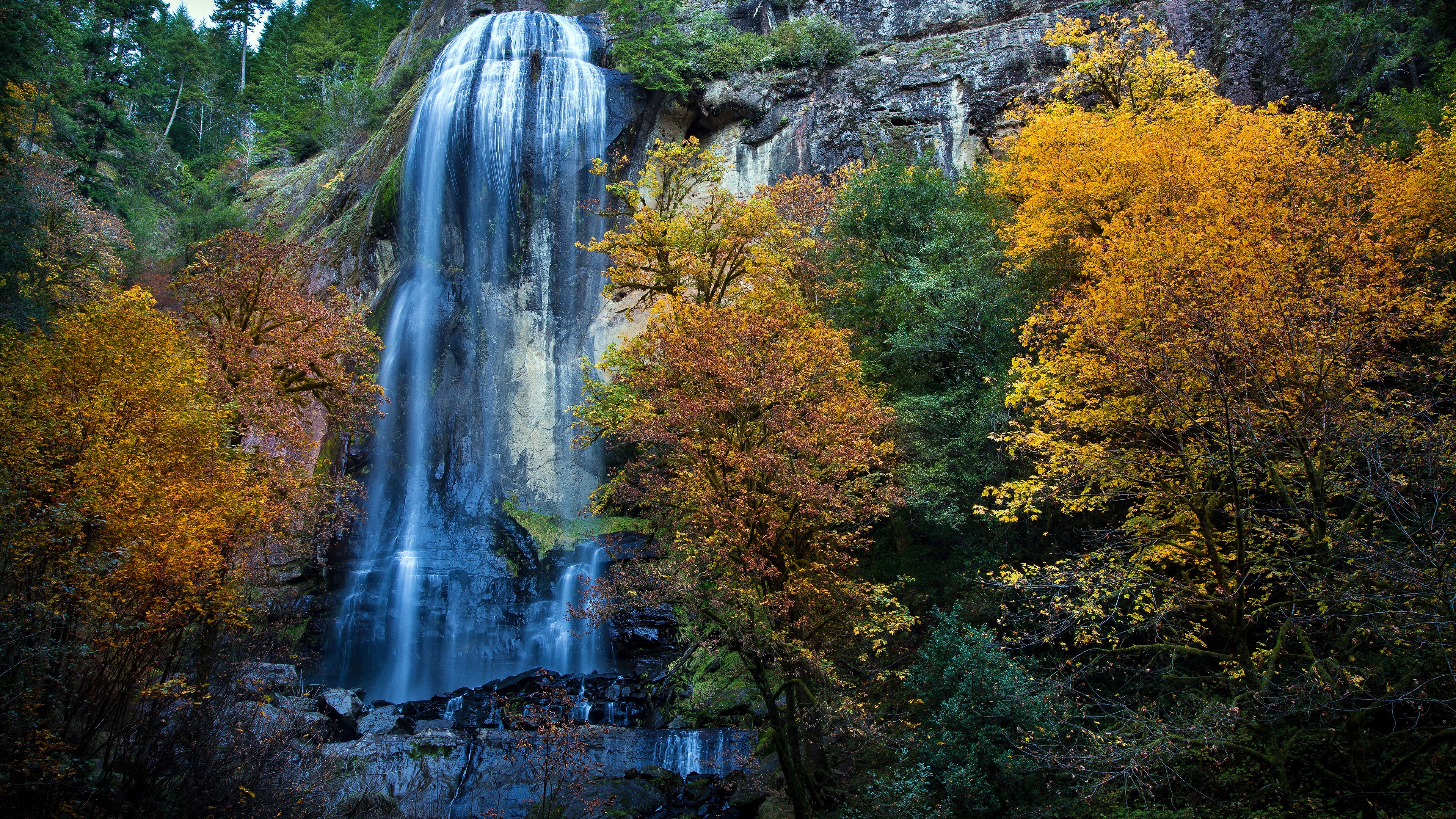 Waterfall cascading down a rocky cliff surrounded by autumn trees HD Wallpaper