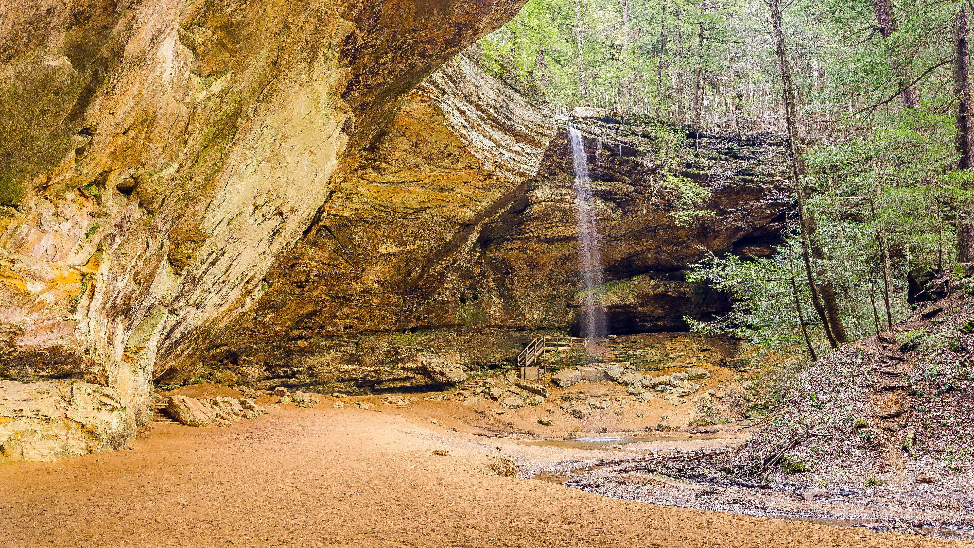Waterfall cascading into a sandy cave with a forest backdrop HD Wallpaper