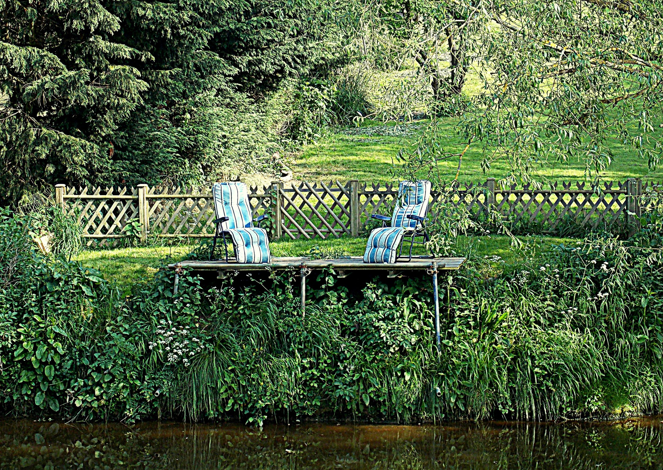 Two chairs on a wooden platform by a fence and river HD Wallpaper