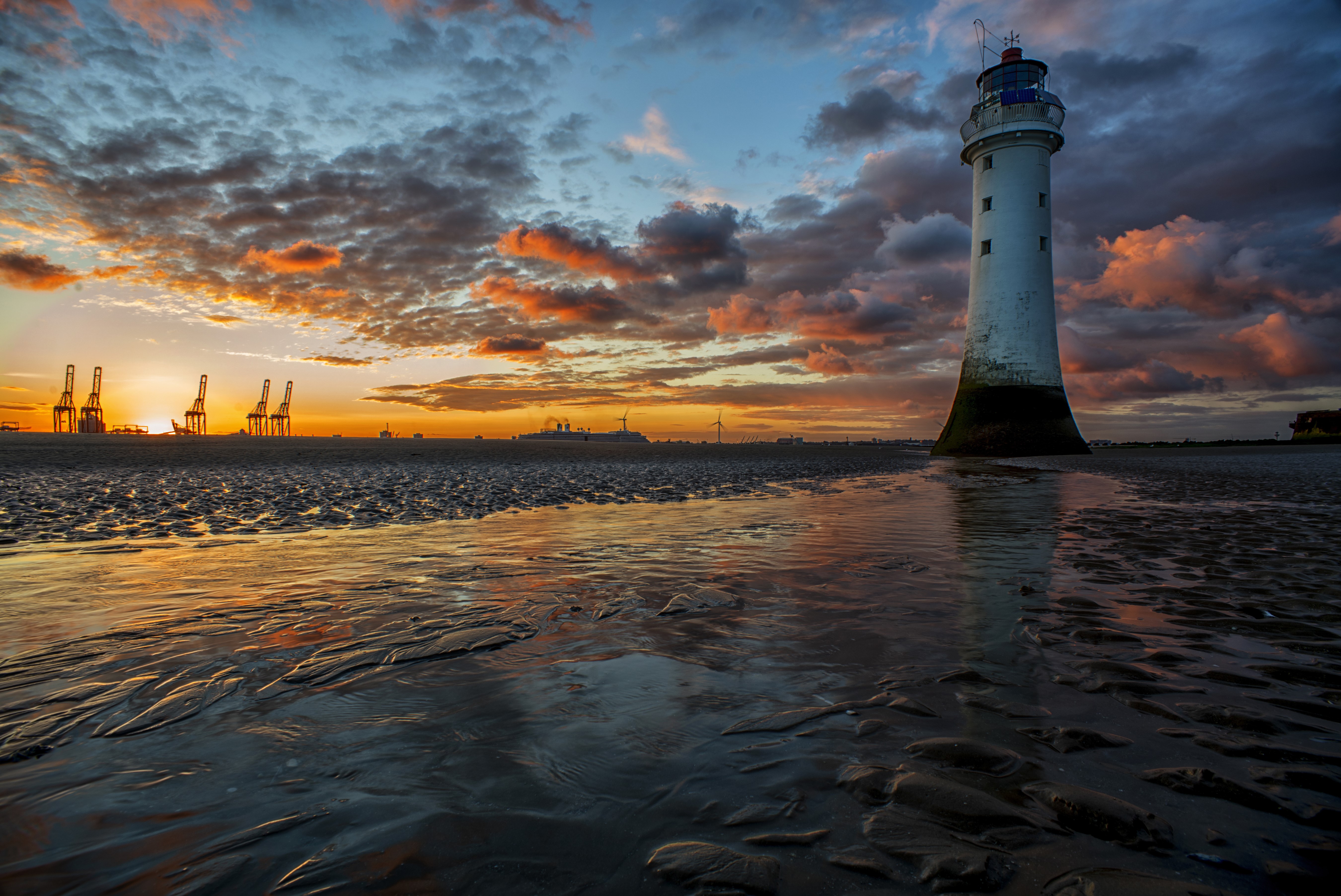 Lighthouse on a beach at sunset with industrial cranes HD Wallpaper