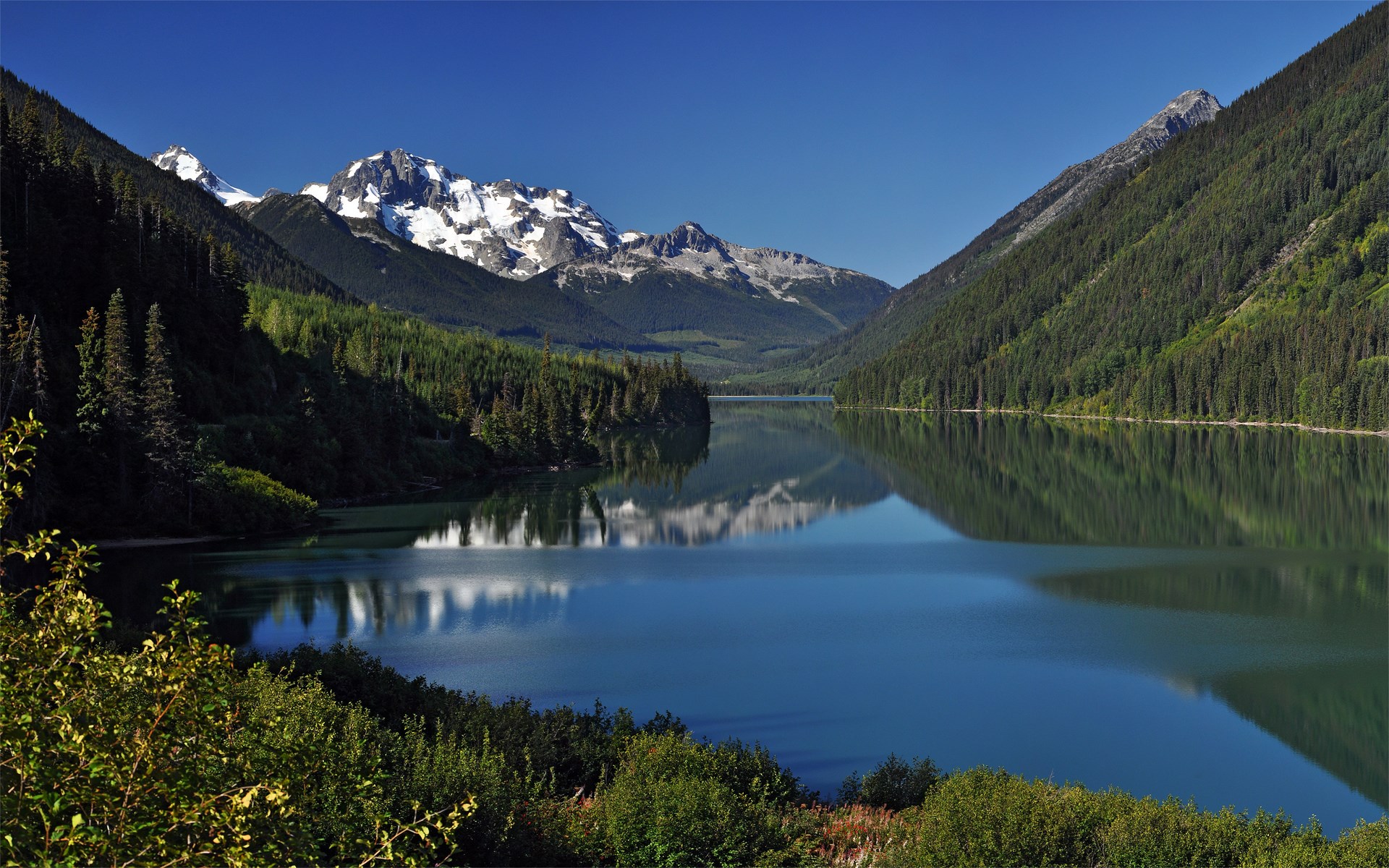 Snow-capped mountains reflected in a calm lake surrounded by forests HD Wallpaper