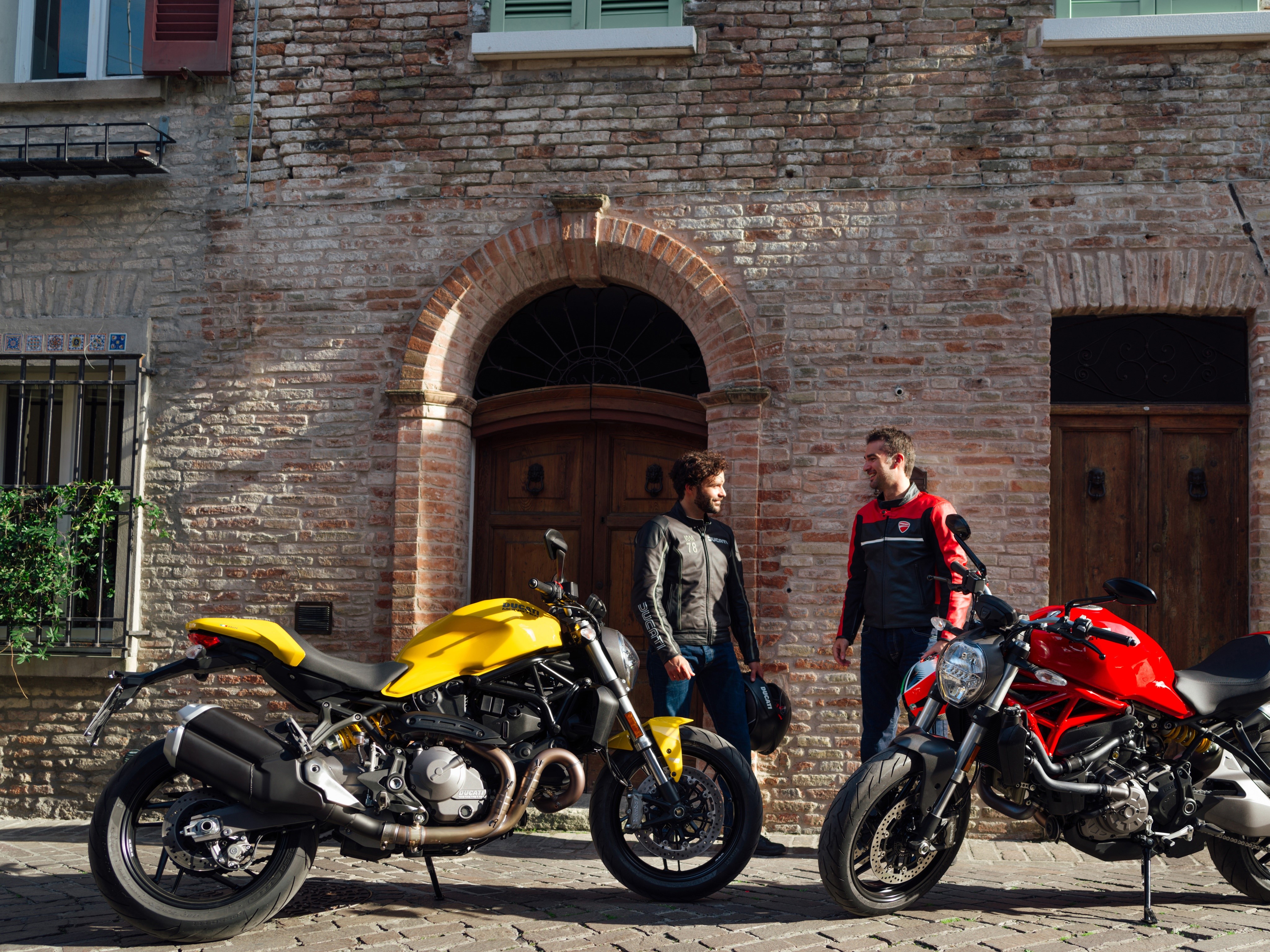 Two men with Ducati motorcycles in front of a brick building HD Wallpaper