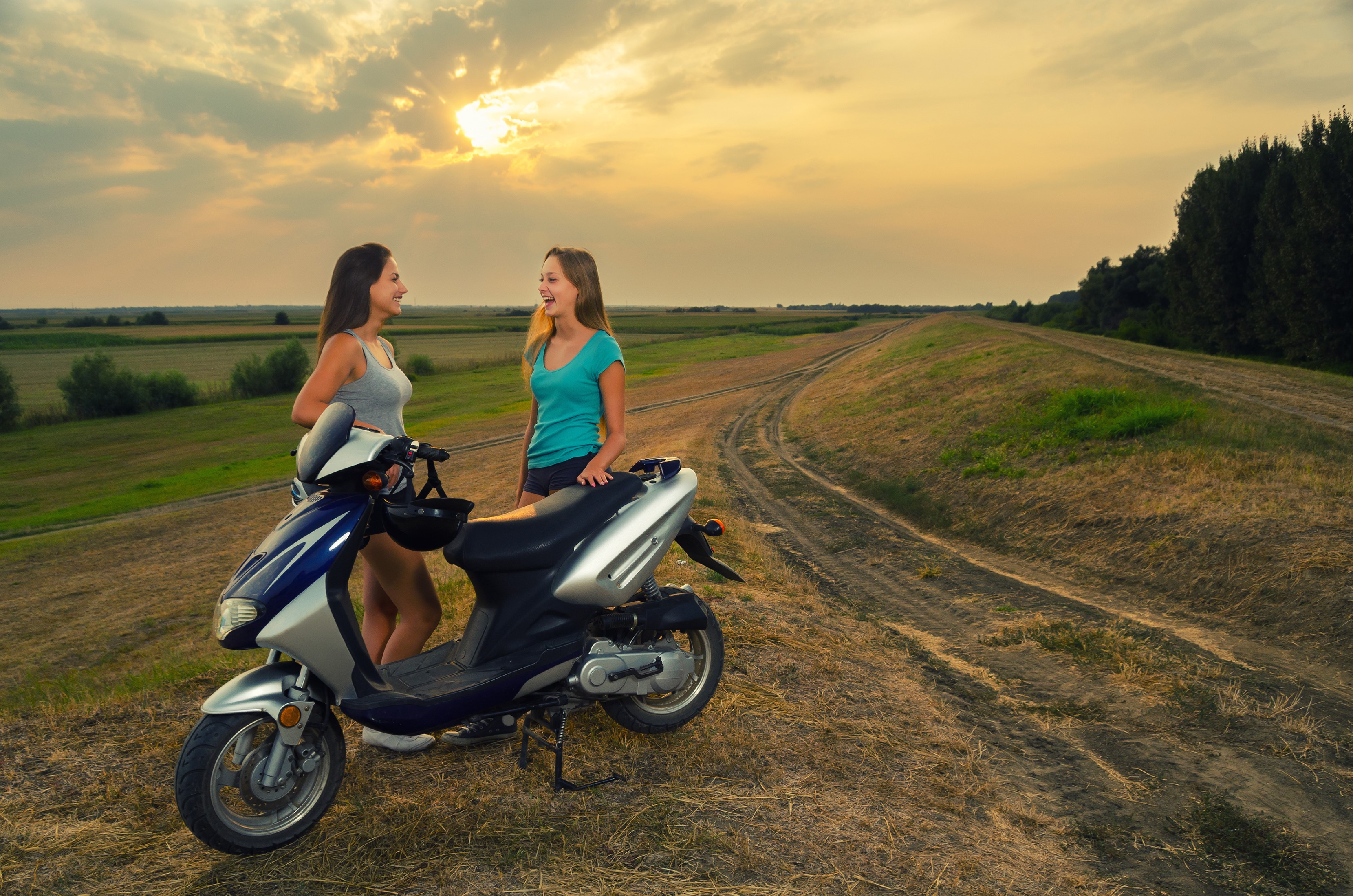Two young women and a scooter on a dirt road at sunset HD Wallpaper