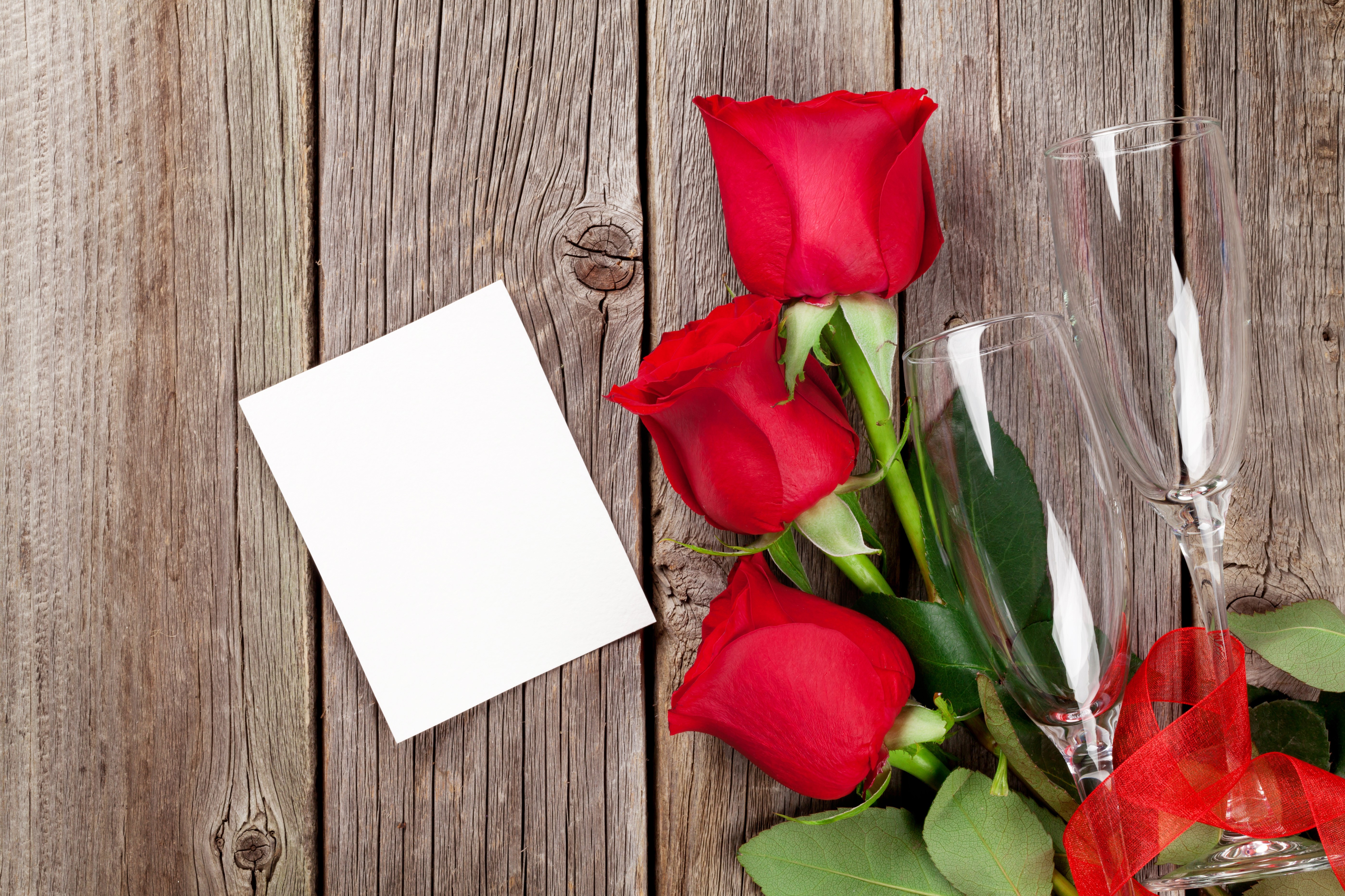 Three red roses with champagne glasses and a card on wood HD Wallpaper