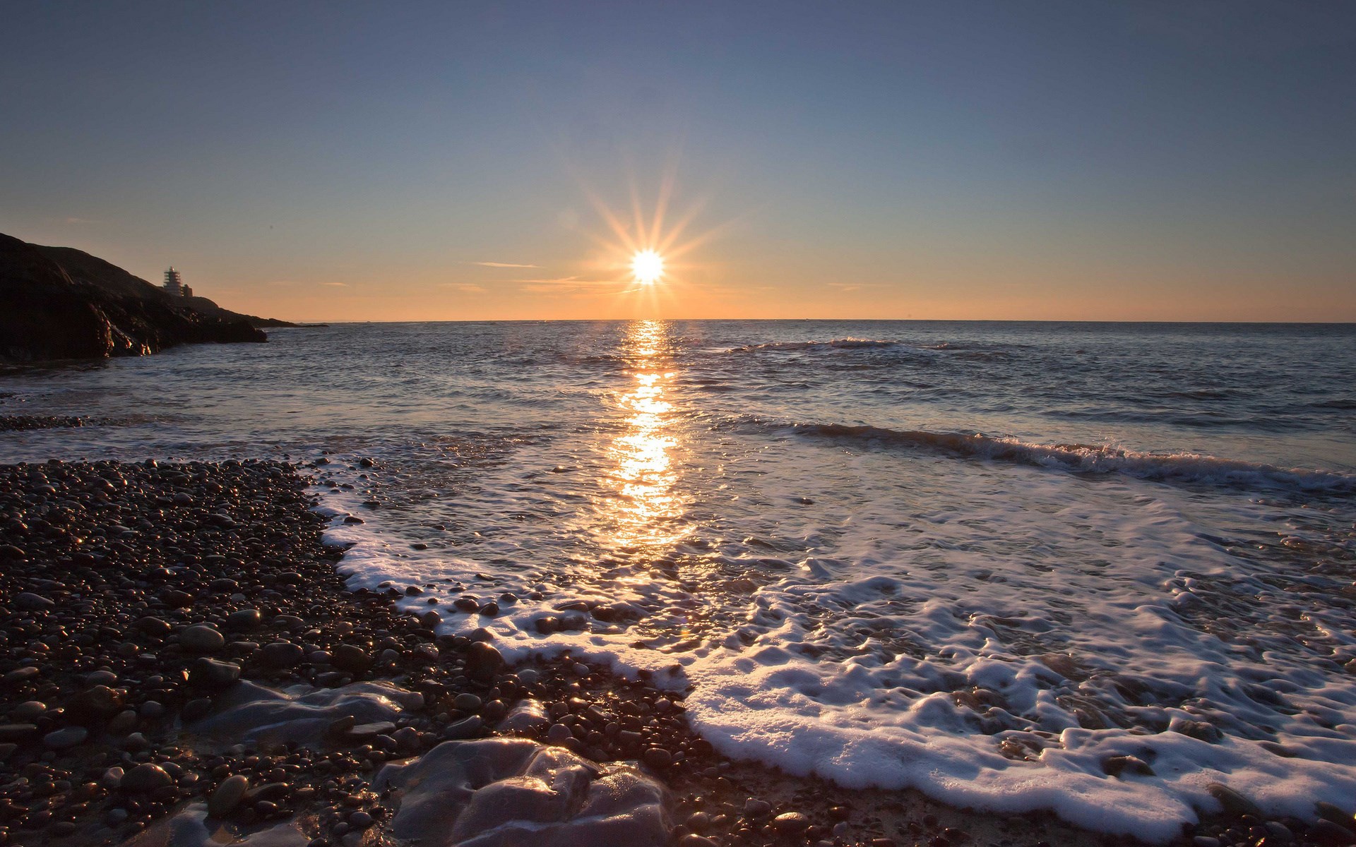 Sunset over the ocean with a lighthouse on a rocky shore HD Wallpaper