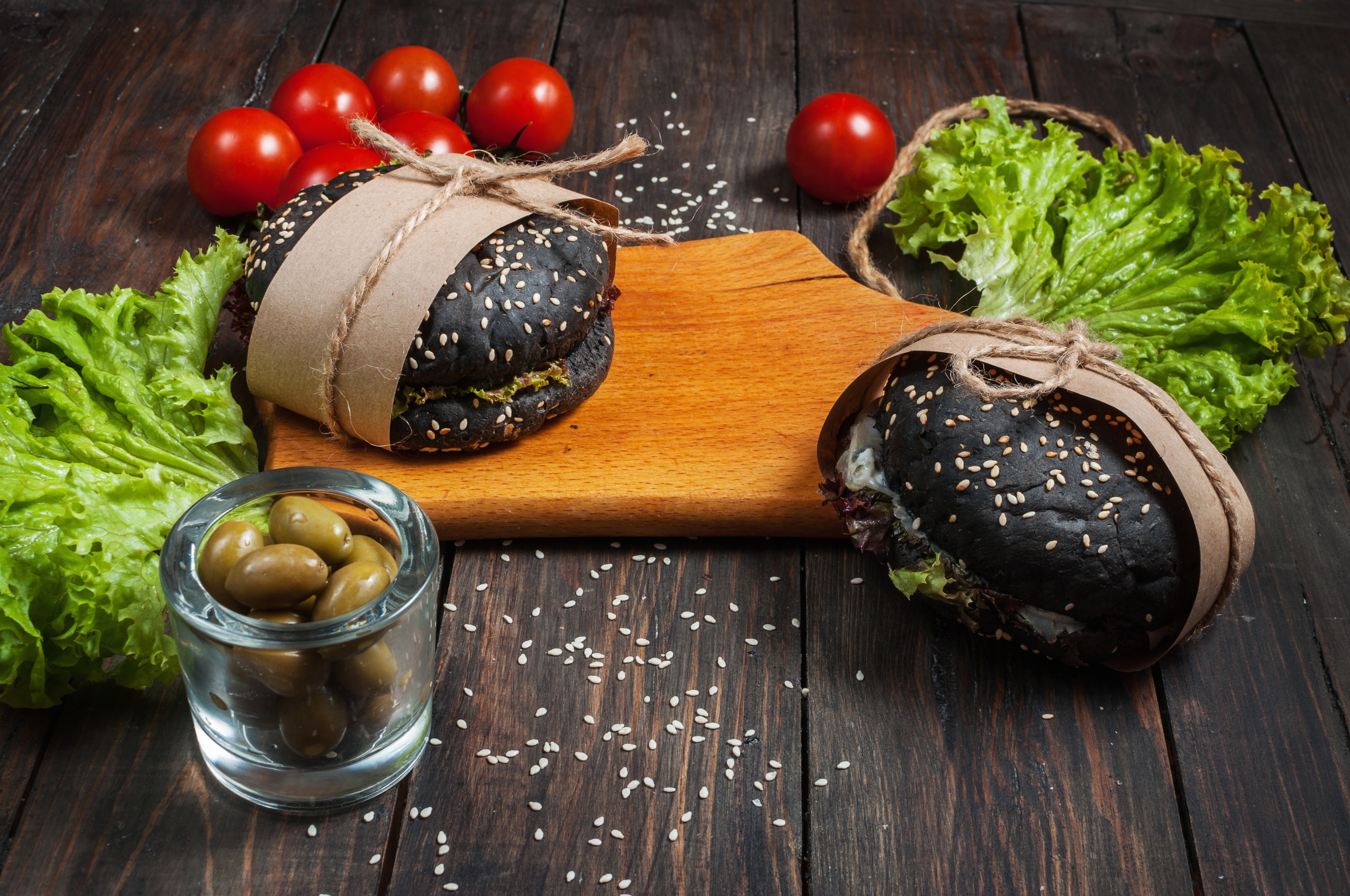 Two black hamburgers with sesame seeds on a wooden cutting board with tomatoes, lettuce, and olives HD Wallpaper