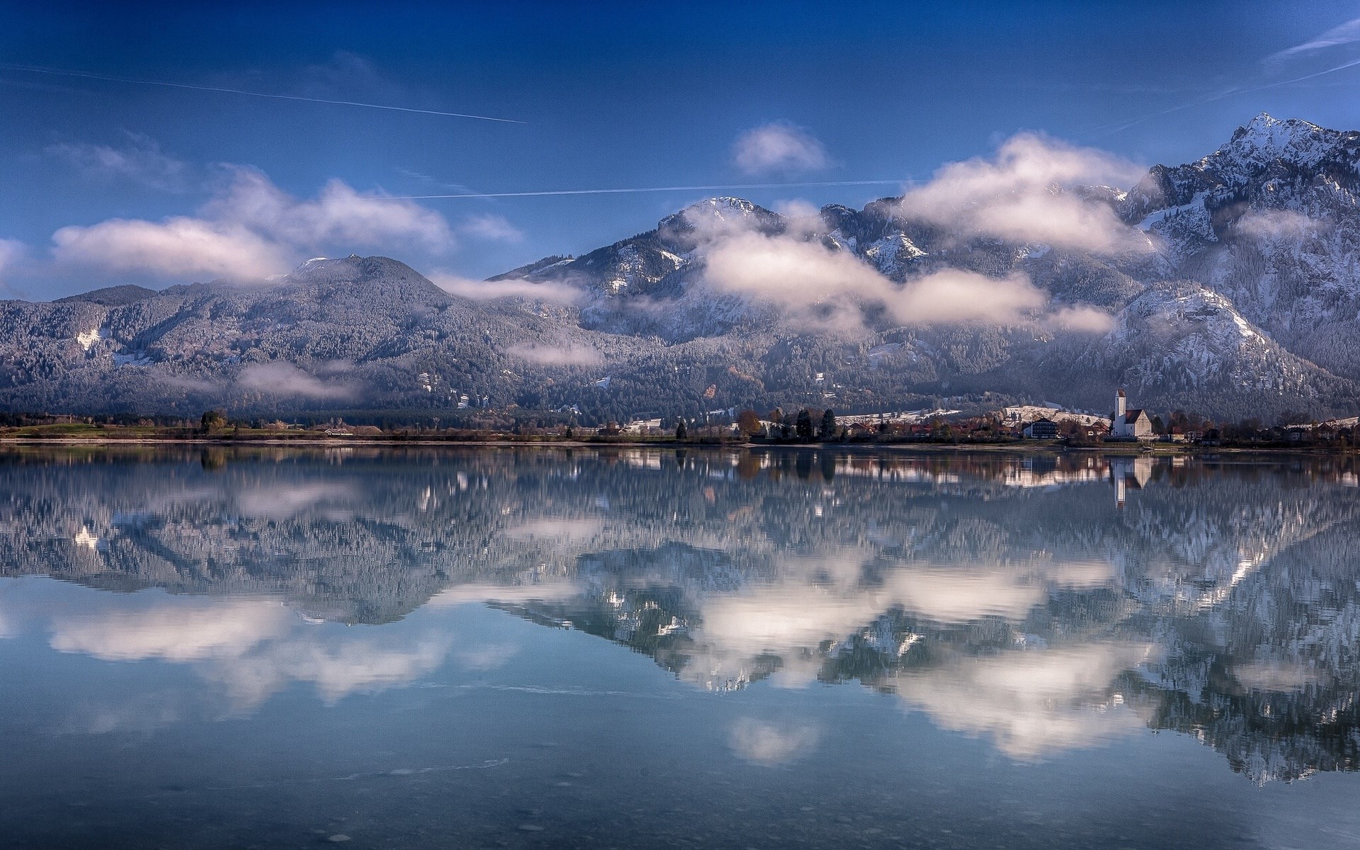 Snow-capped mountains reflected in a calm lake with a village HD Wallpaper