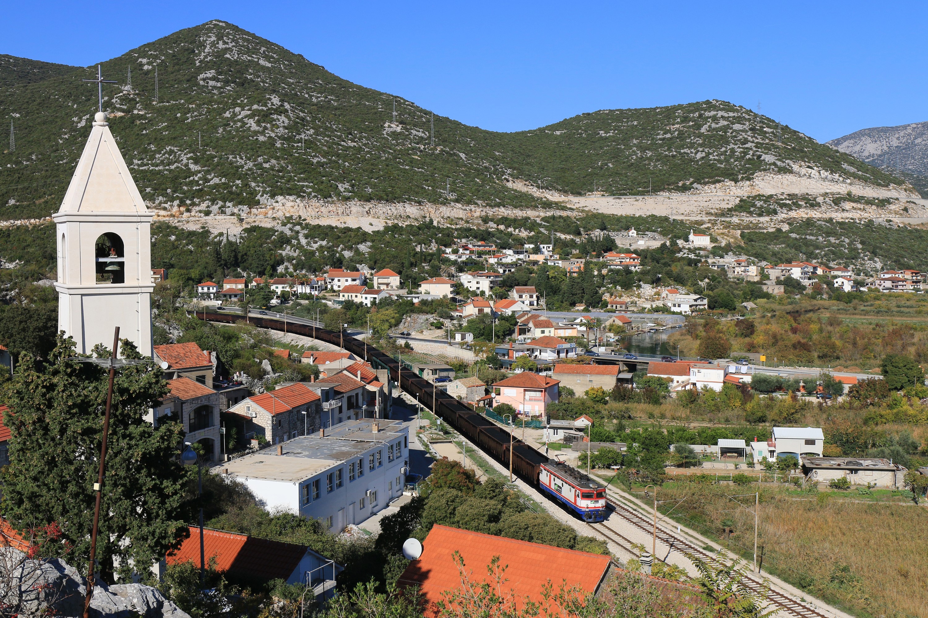 Train passing through a village with a church tower and mountains HD Wallpaper