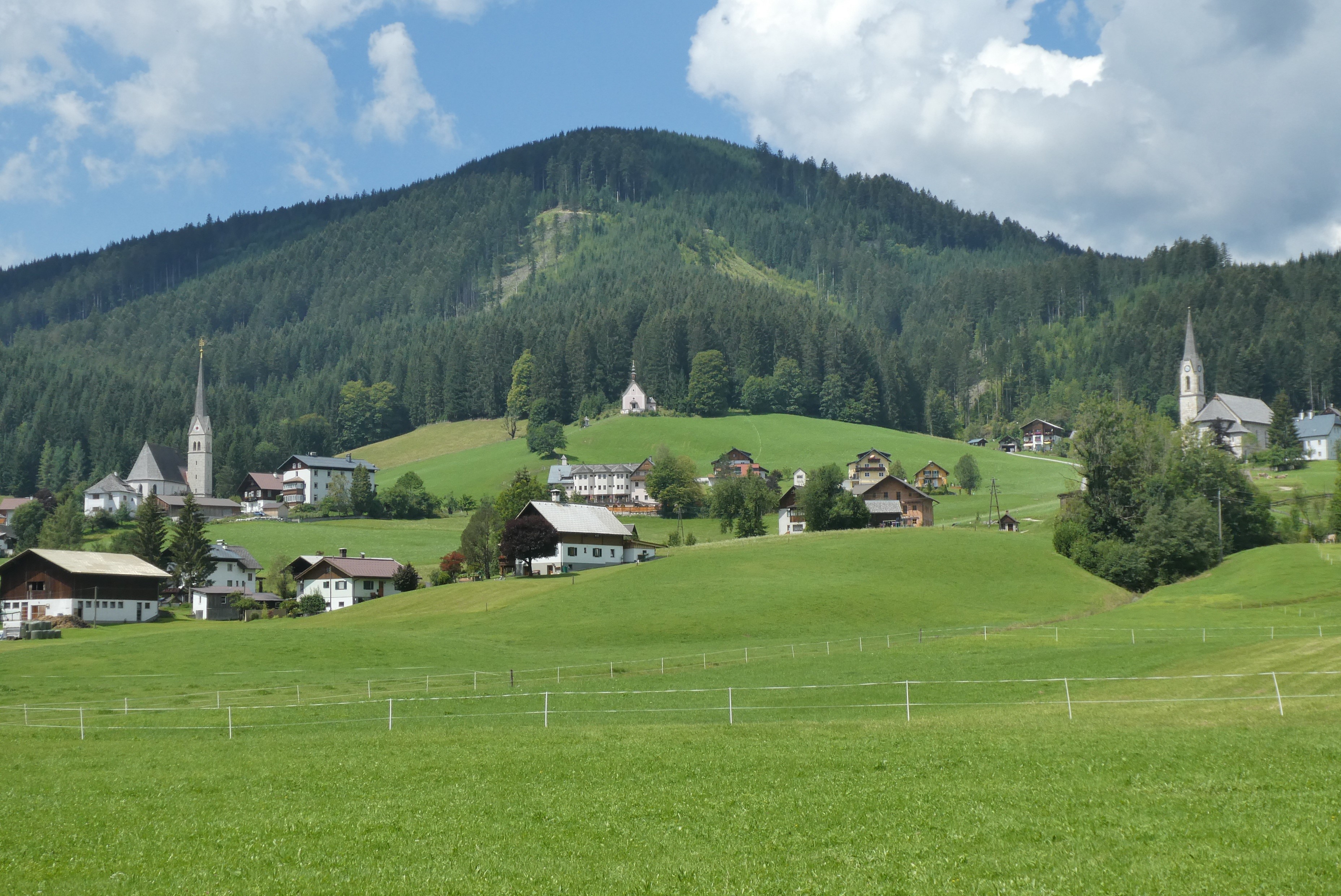 Alpine village with churches and forested mountain HD Wallpaper