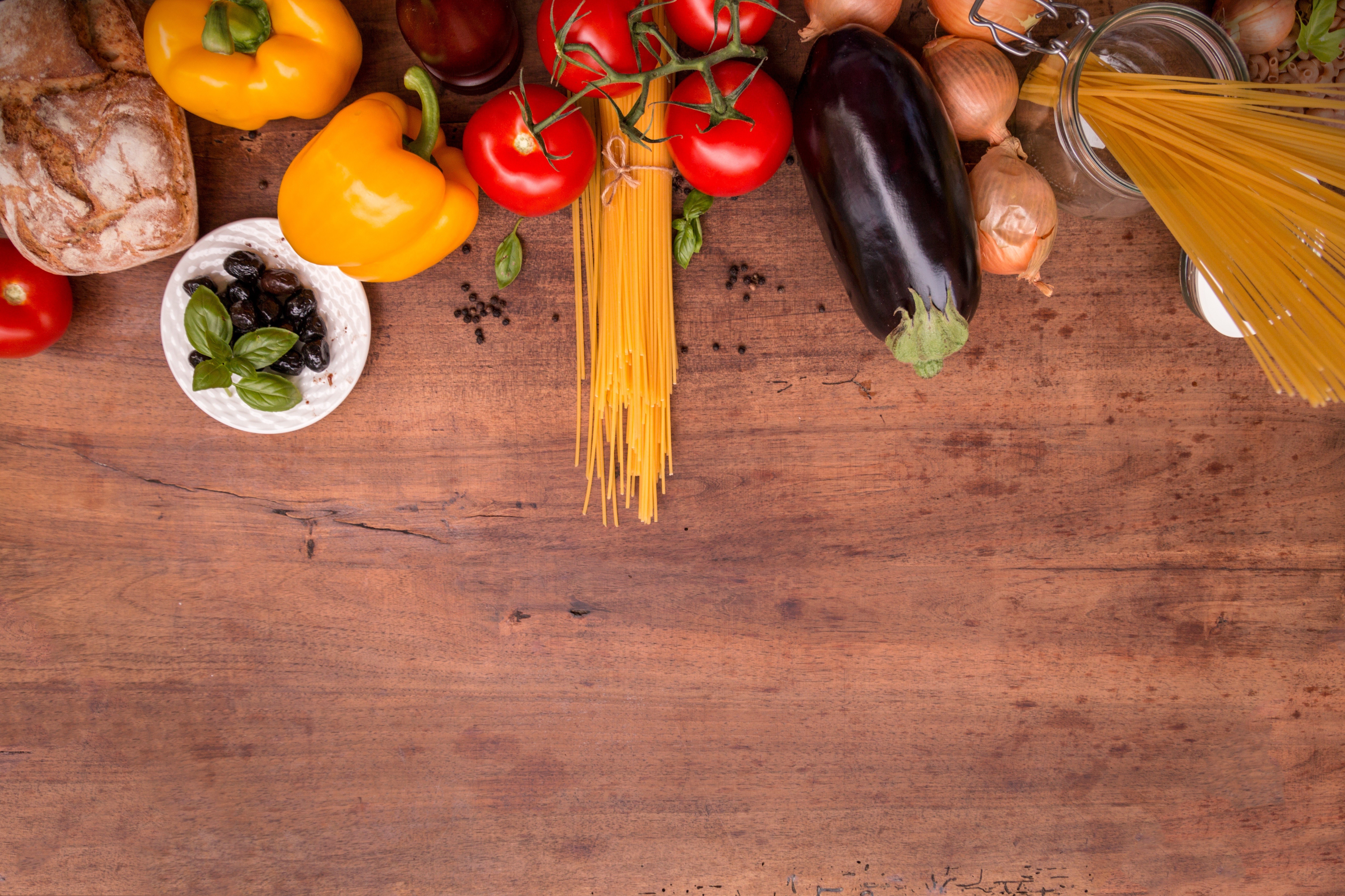 Bell peppers, tomatoes, eggplant, onions, pasta, olives, and bread on a wooden table HD Wallpaper