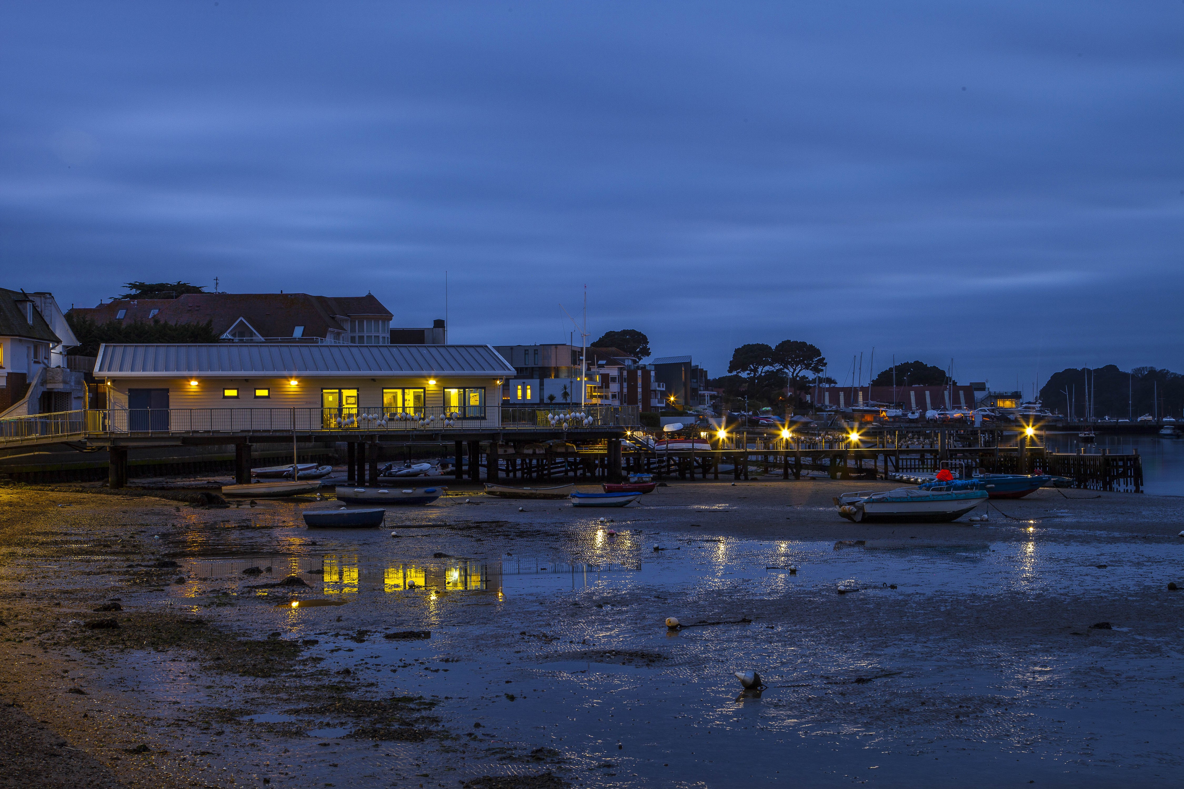 Boats on a muddy shore at dusk with buildings and lights HD Wallpaper