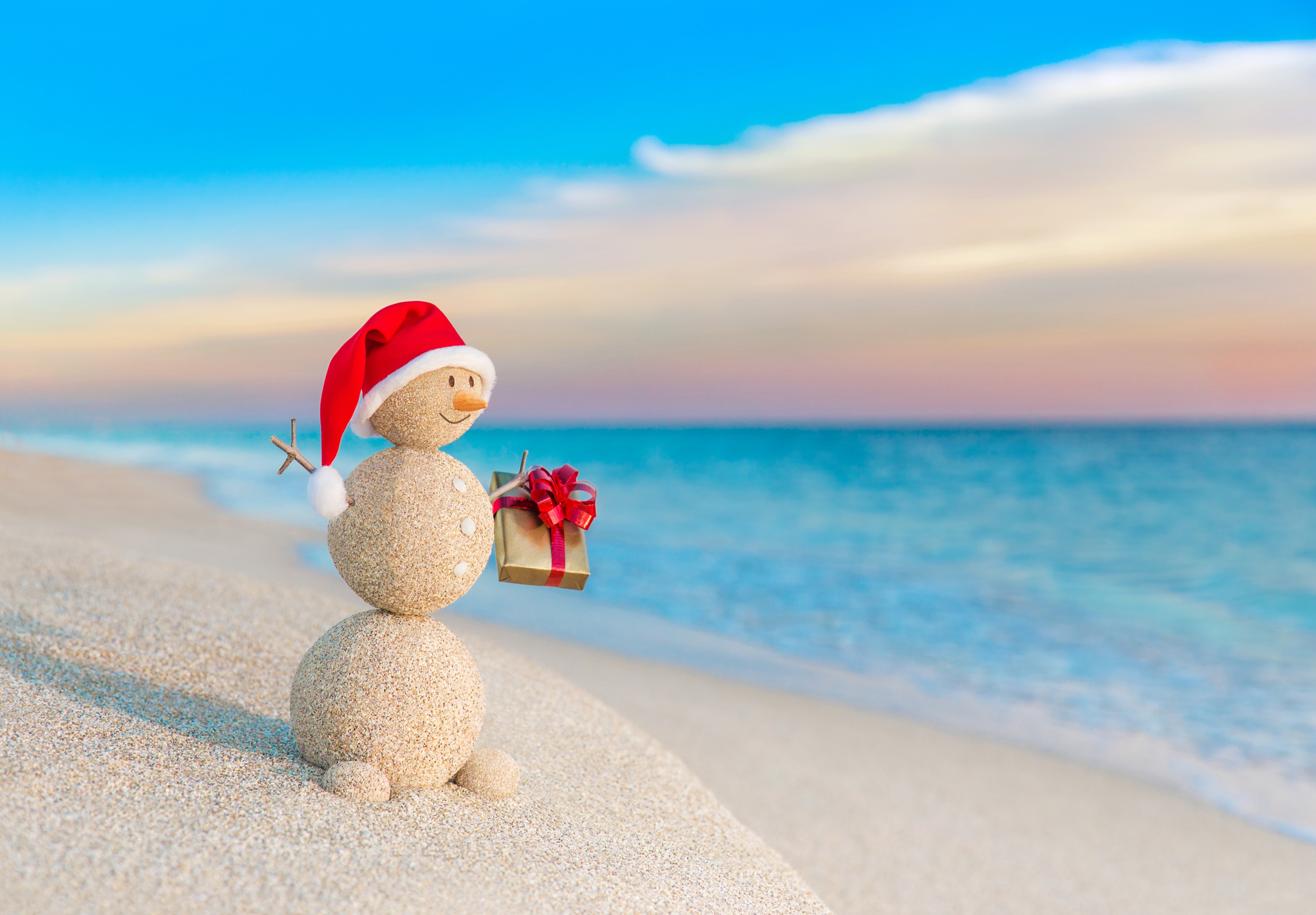 Sand snowman with Christmas hat and gift on beach HD Wallpaper