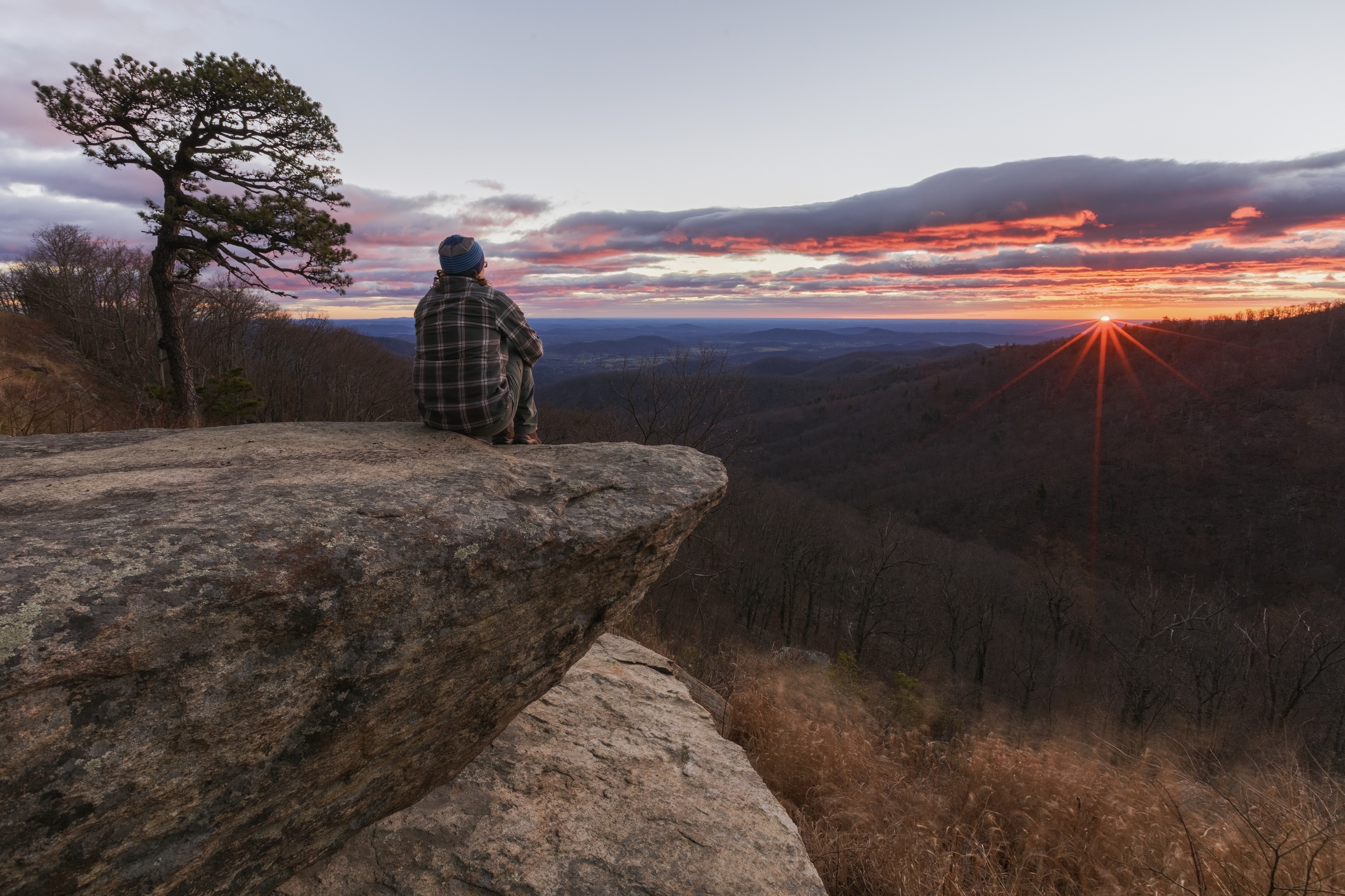 Person sitting on a rock overlooking a sunset in the mountains HD Wallpaper
