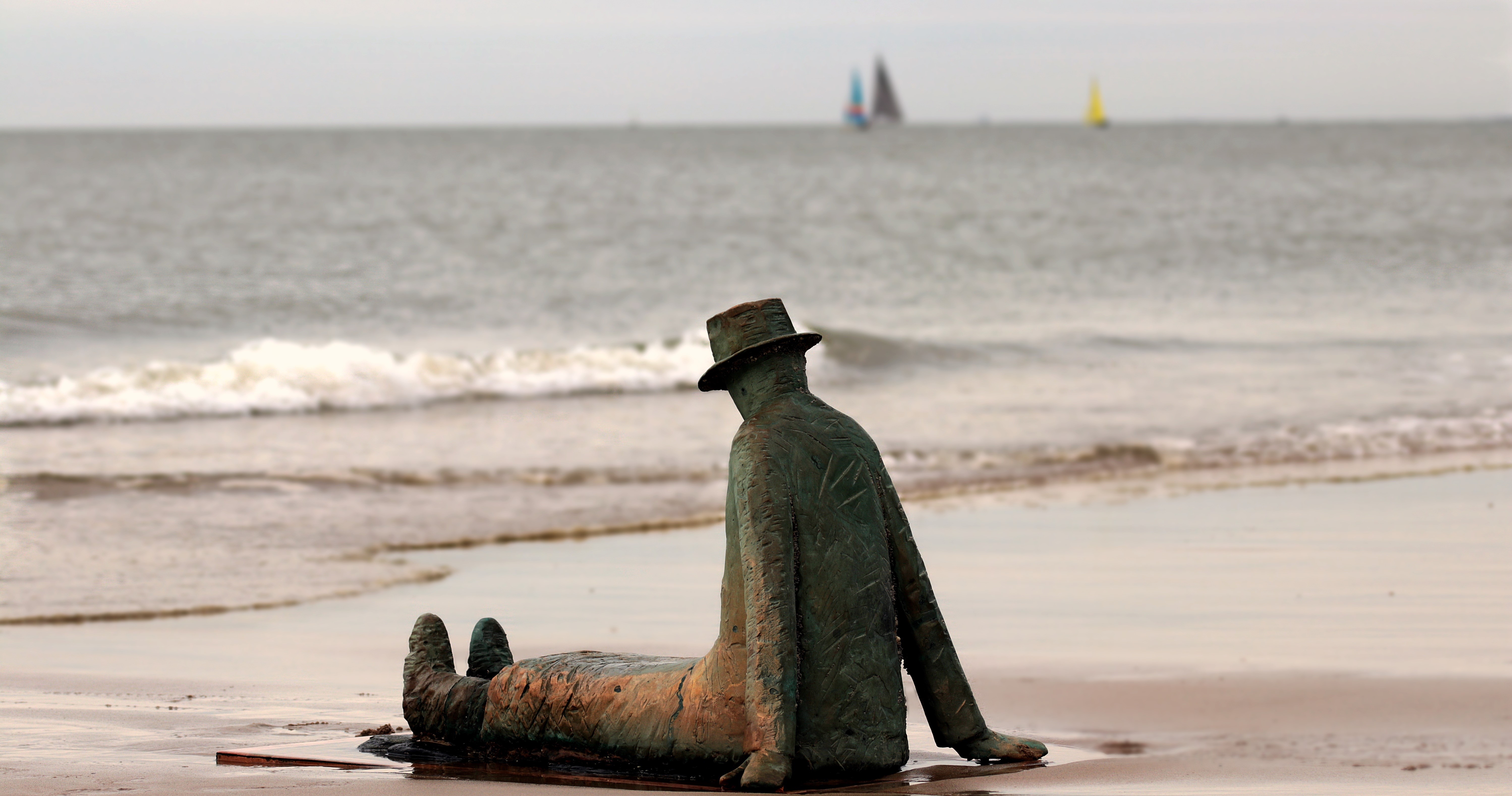 Sculpture of a man in a hat sitting on a beach HD Wallpaper