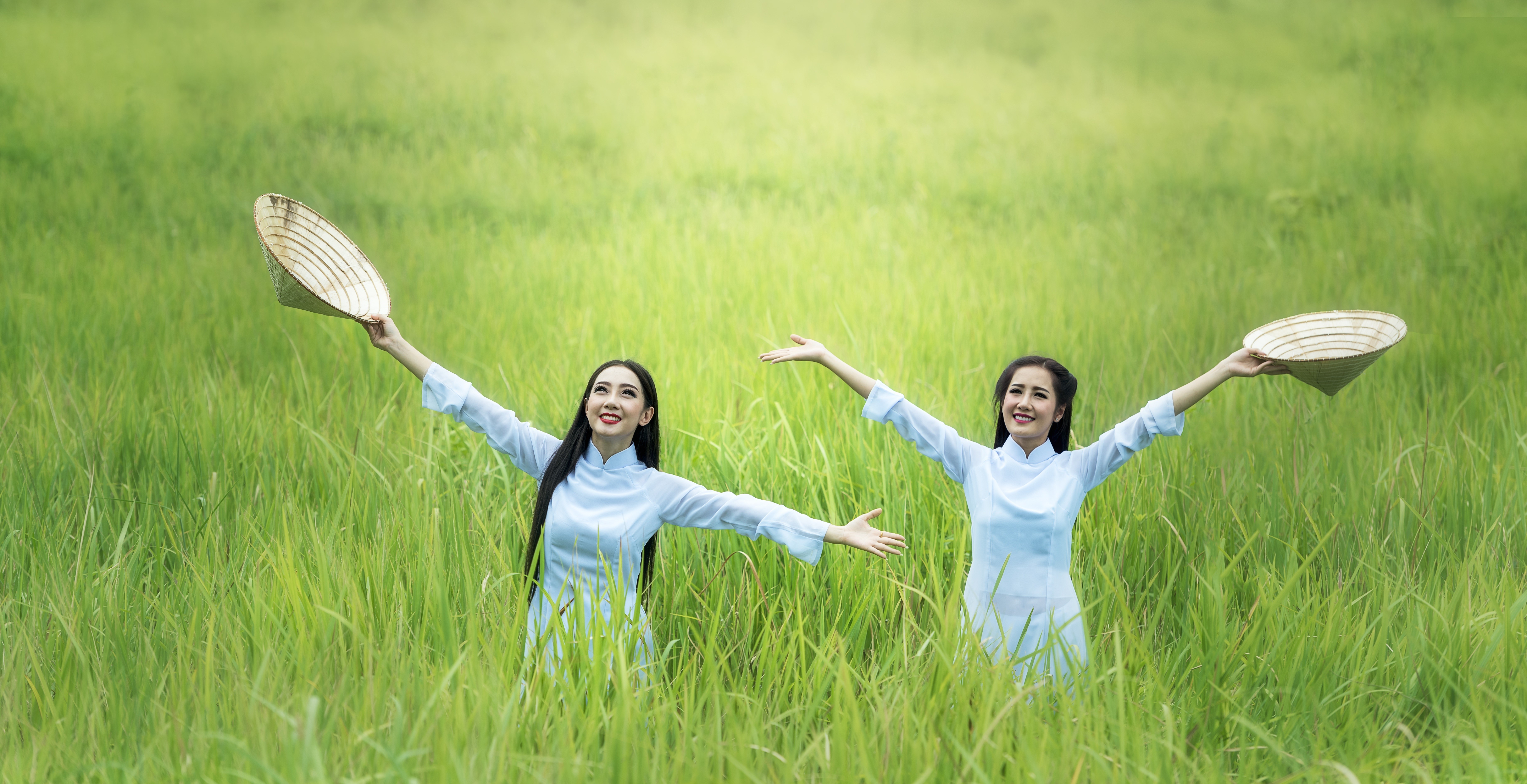 Two Asian women in white dresses holding hats in a grassy field HD Wallpaper