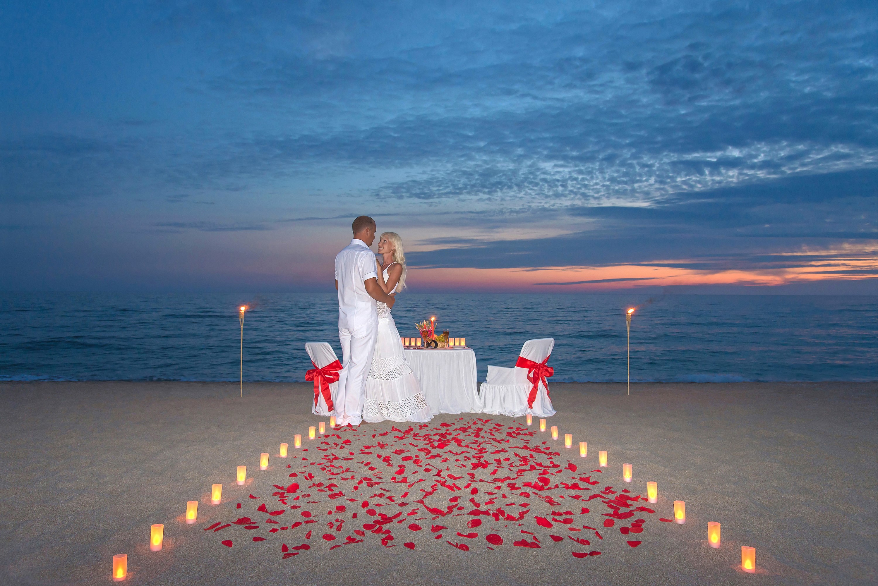 Couple embracing on a beach at sunset with candles and rose petals HD Wallpaper