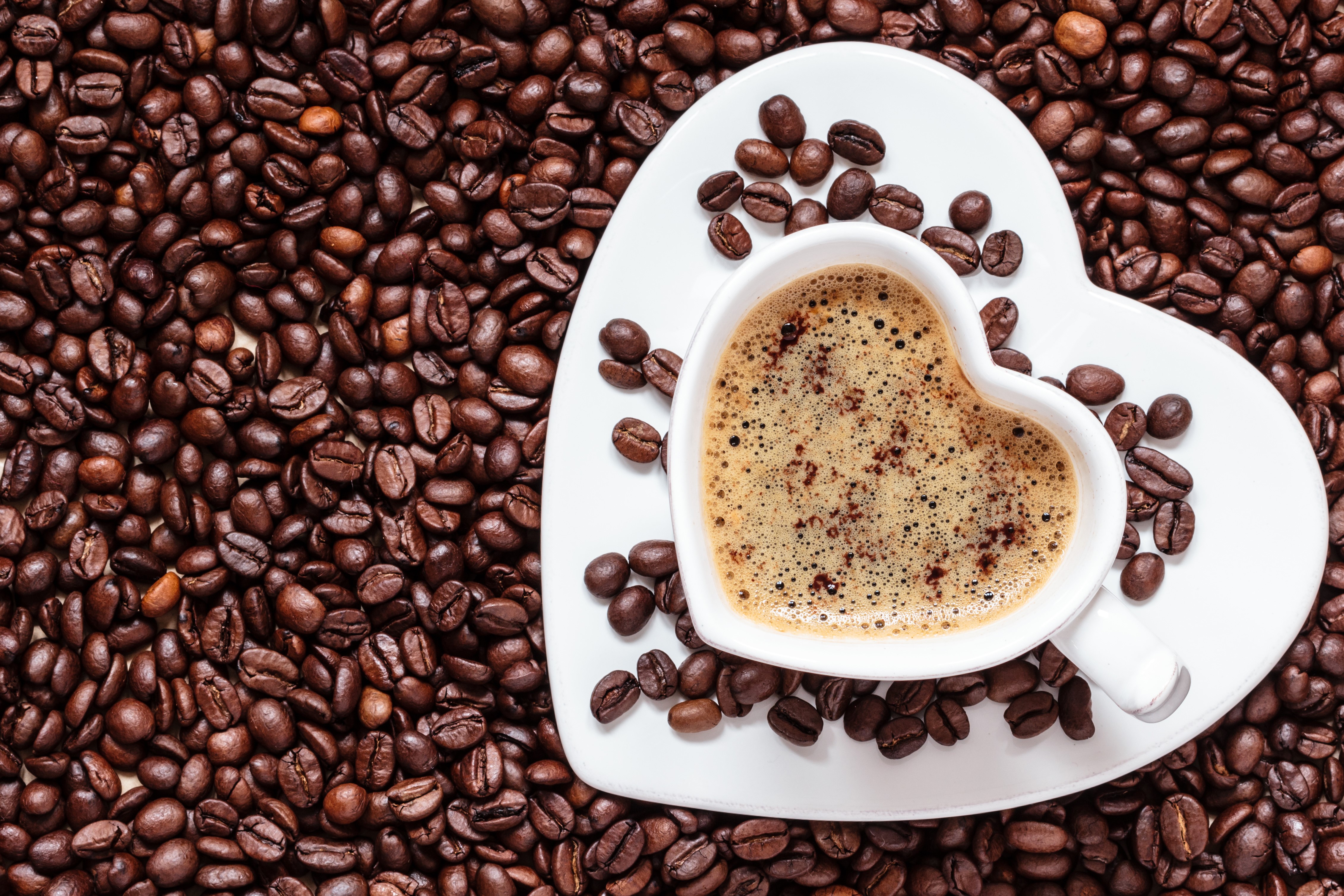 Heart-shaped cup of coffee on a saucer surrounded by coffee beans HD Wallpaper